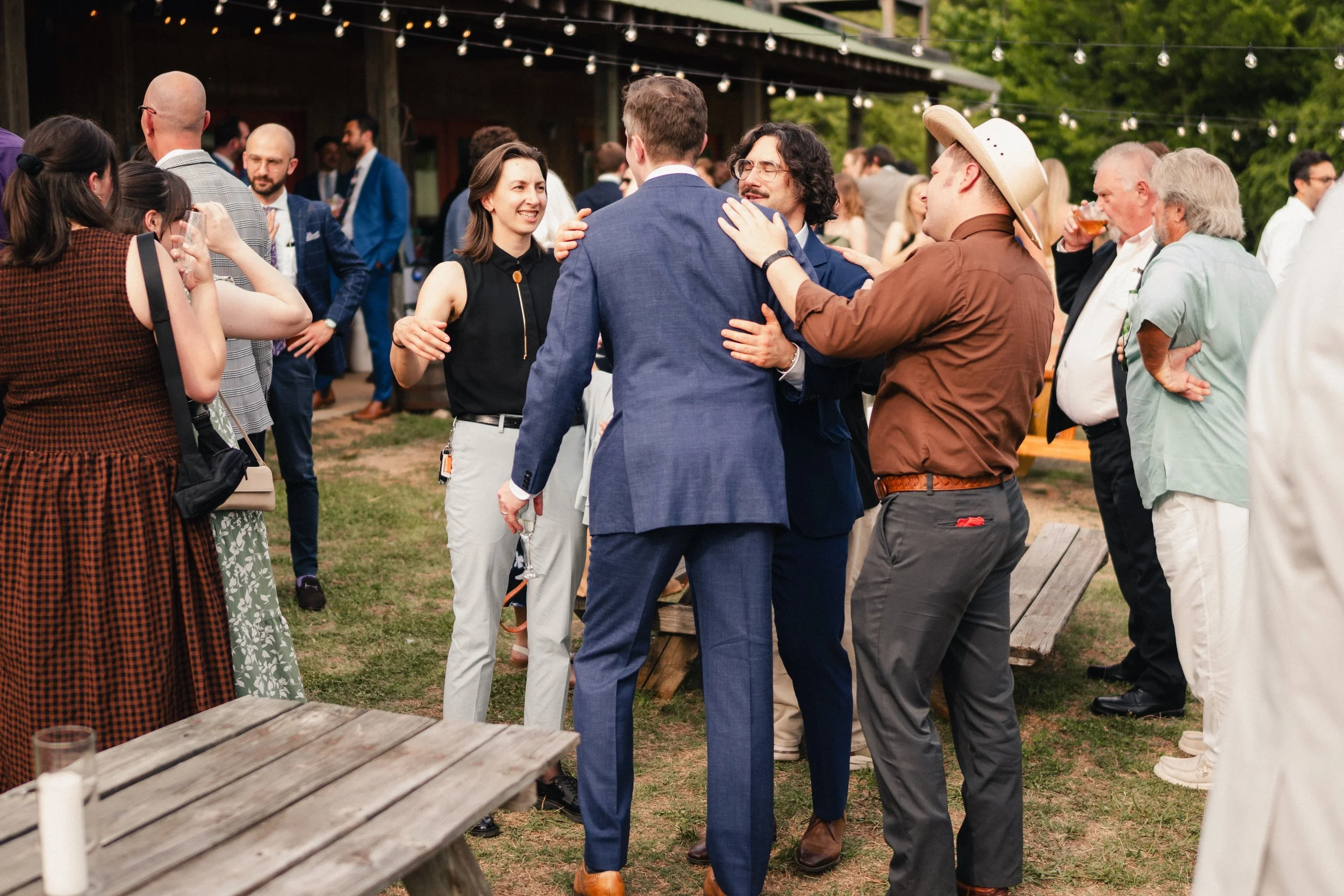 Group of people socializing at an outdoor party or wedding reception under string lights, with some holding drinks and engaging in conversations.