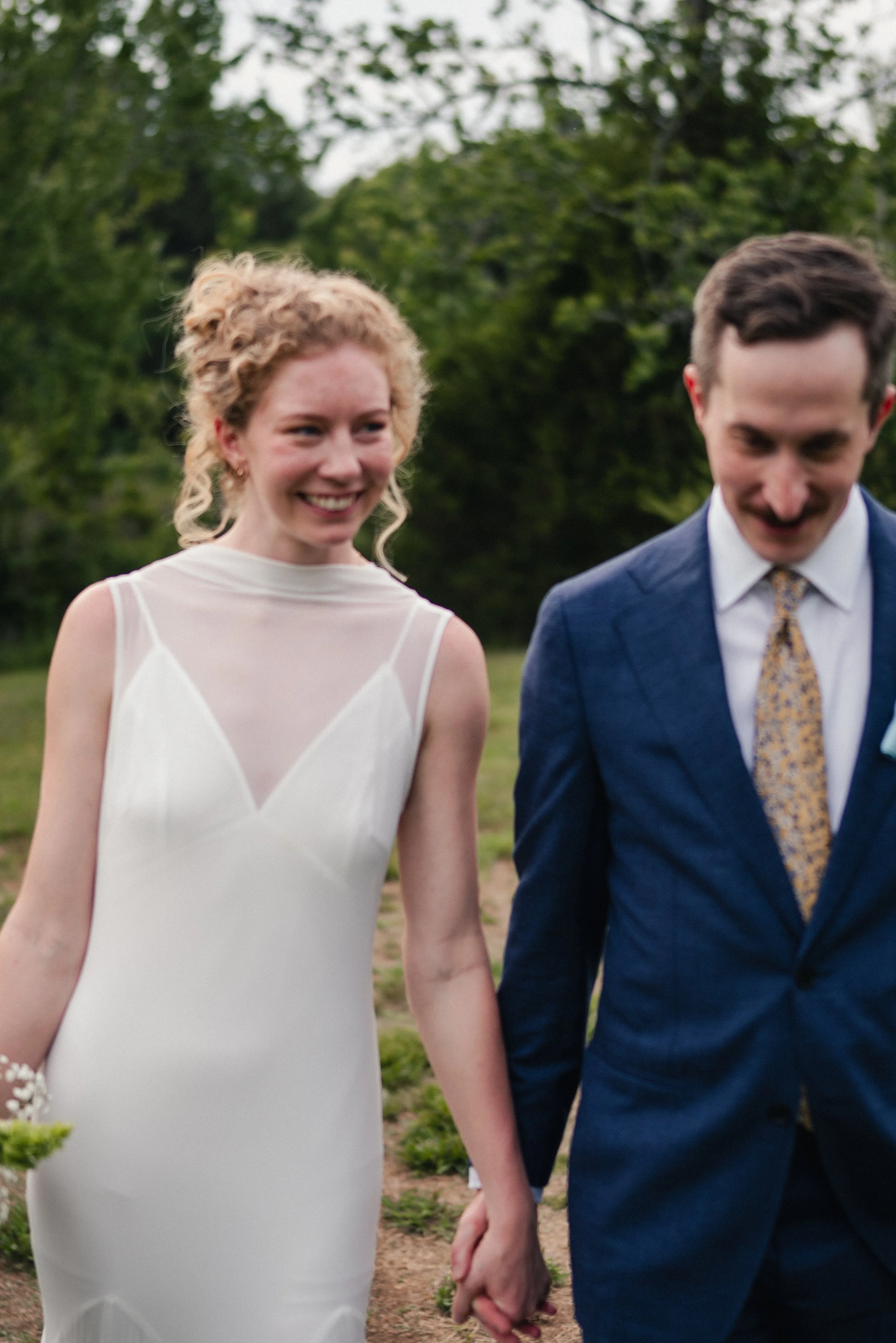 A smiling woman in a white dress and a man in a suit holding hands during an outdoor wedding ceremony with trees in the background.