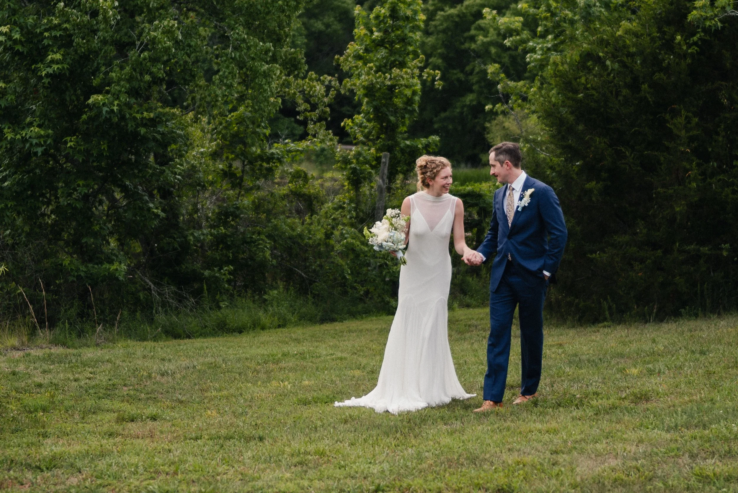 A bride and groom holding hands and smiling at each other outdoors in a green, wooded area.