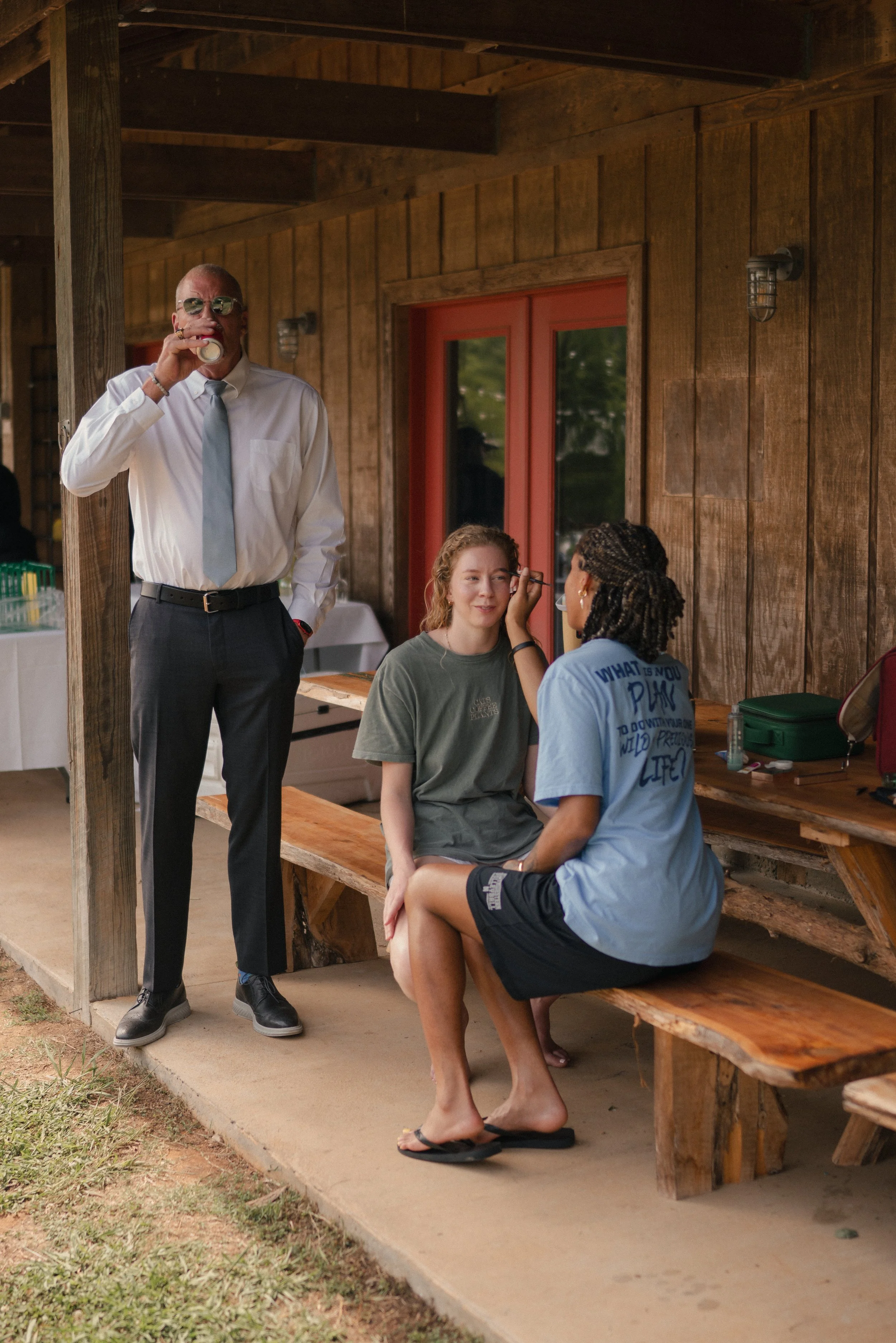 A man in a white shirt, tie, and sunglasses is standing and drinking from a can. Two women are sitting on a wooden bench; one woman is applying makeup to the other, who is smiling. They are in a rustic outdoor setting with wooden walls and a door wit