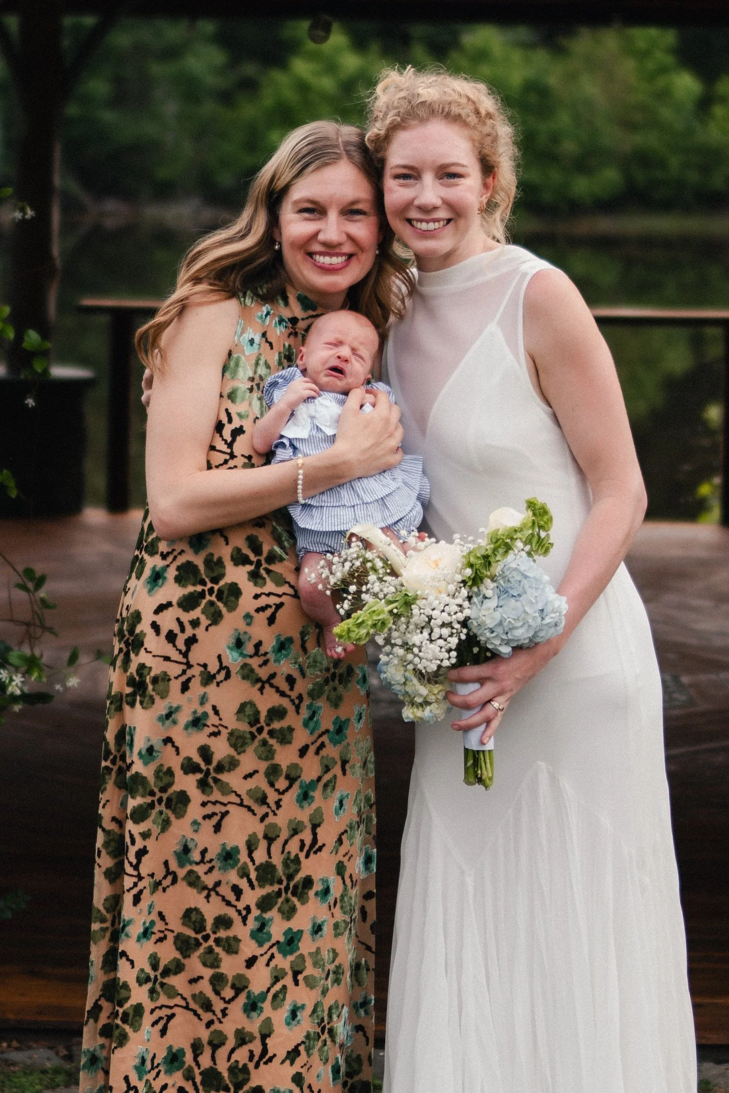 Two women, one holding a crying baby, standing outdoors with greenery in the background. The woman on the left is wearing a floral dress, and the woman on the right is dressed in a white gown holding a bouquet of flowers.