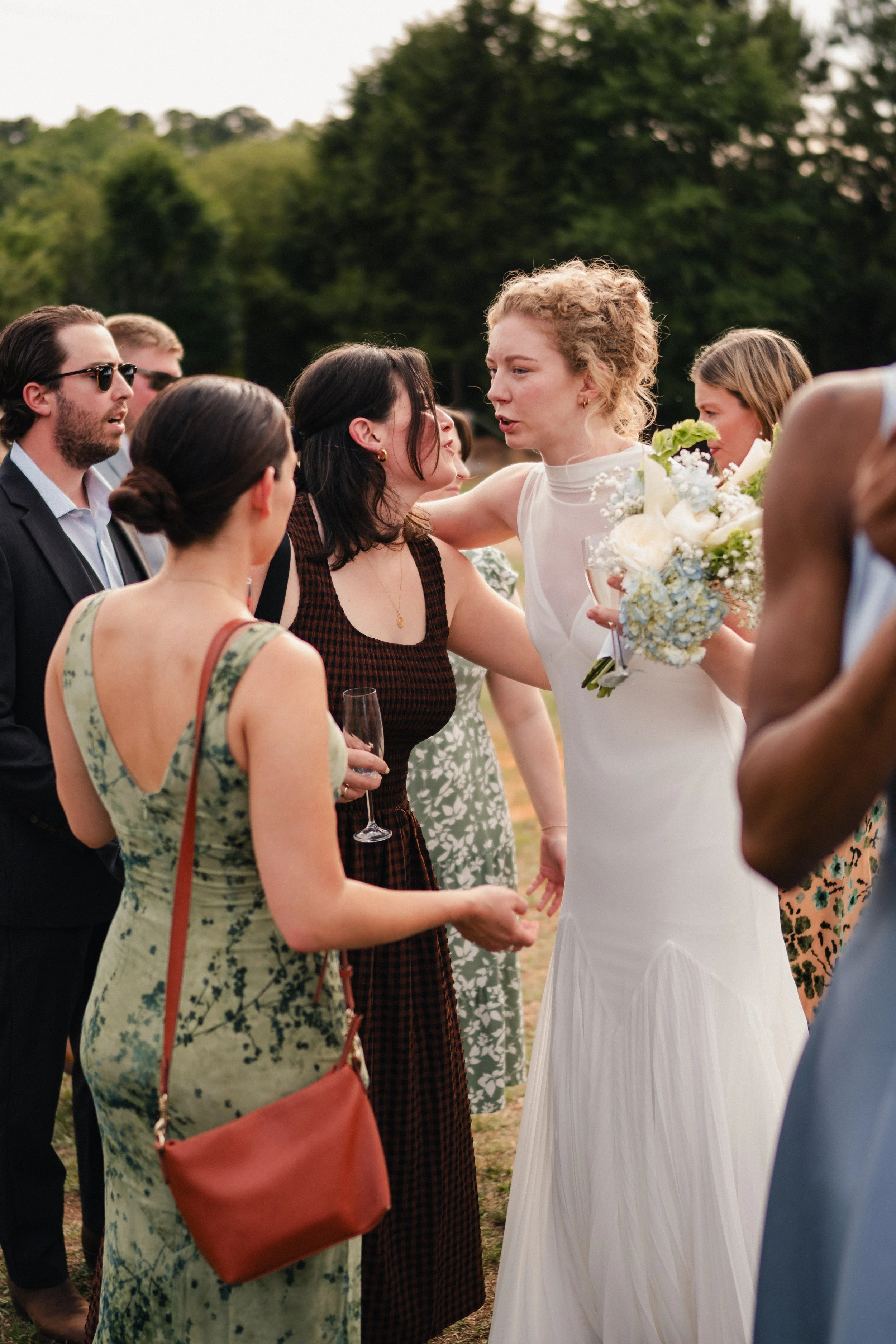 Group of people gathered outdoors, engaged in a conversation. A woman in a white dress holding a bouquet of flowers is at the center.
