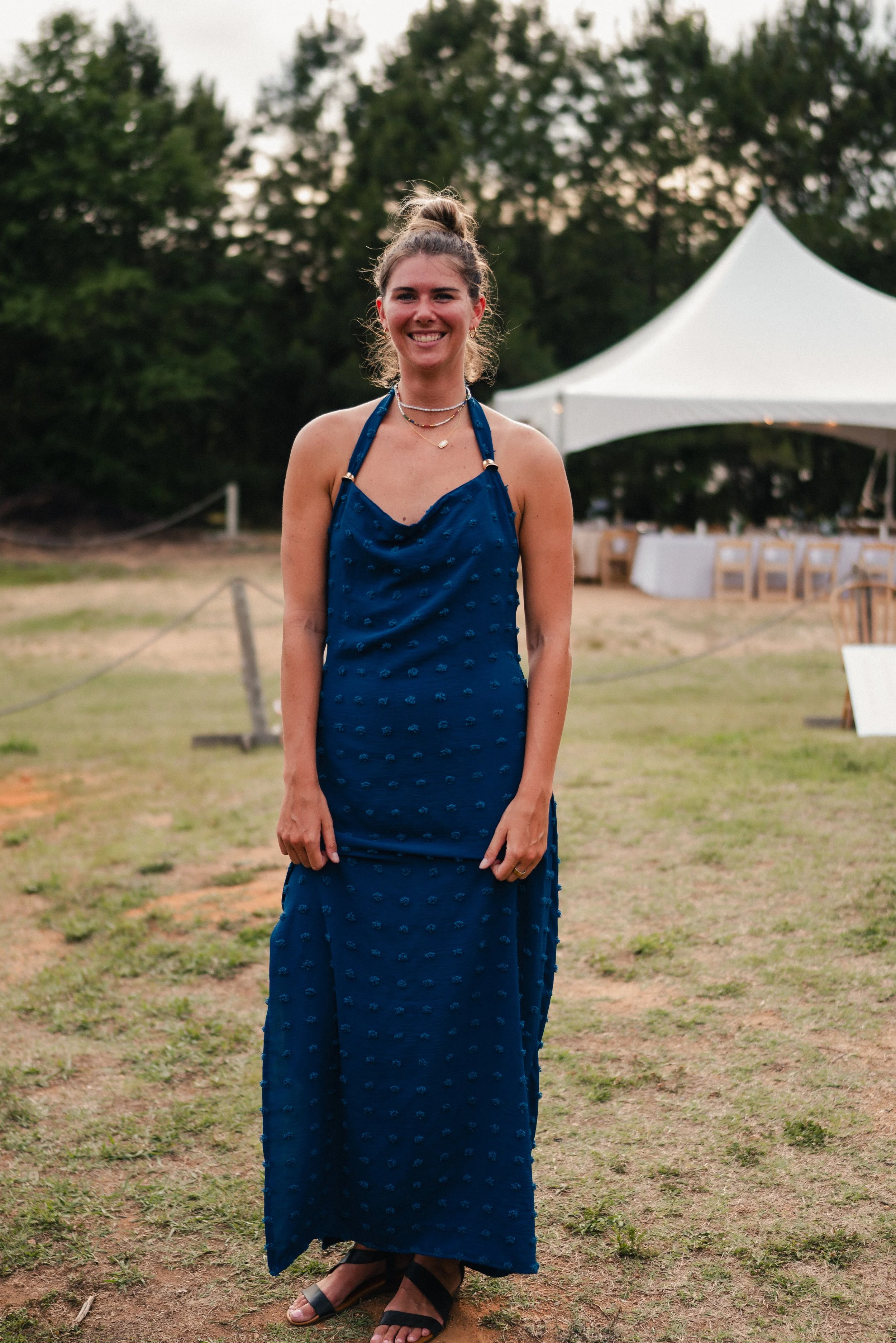 A young woman with light skin, smiling, wearing a blue dress with small floral patterns, layered necklaces, and black sandals, standing outdoors at a daytime event with a large white tent and green trees in the background.