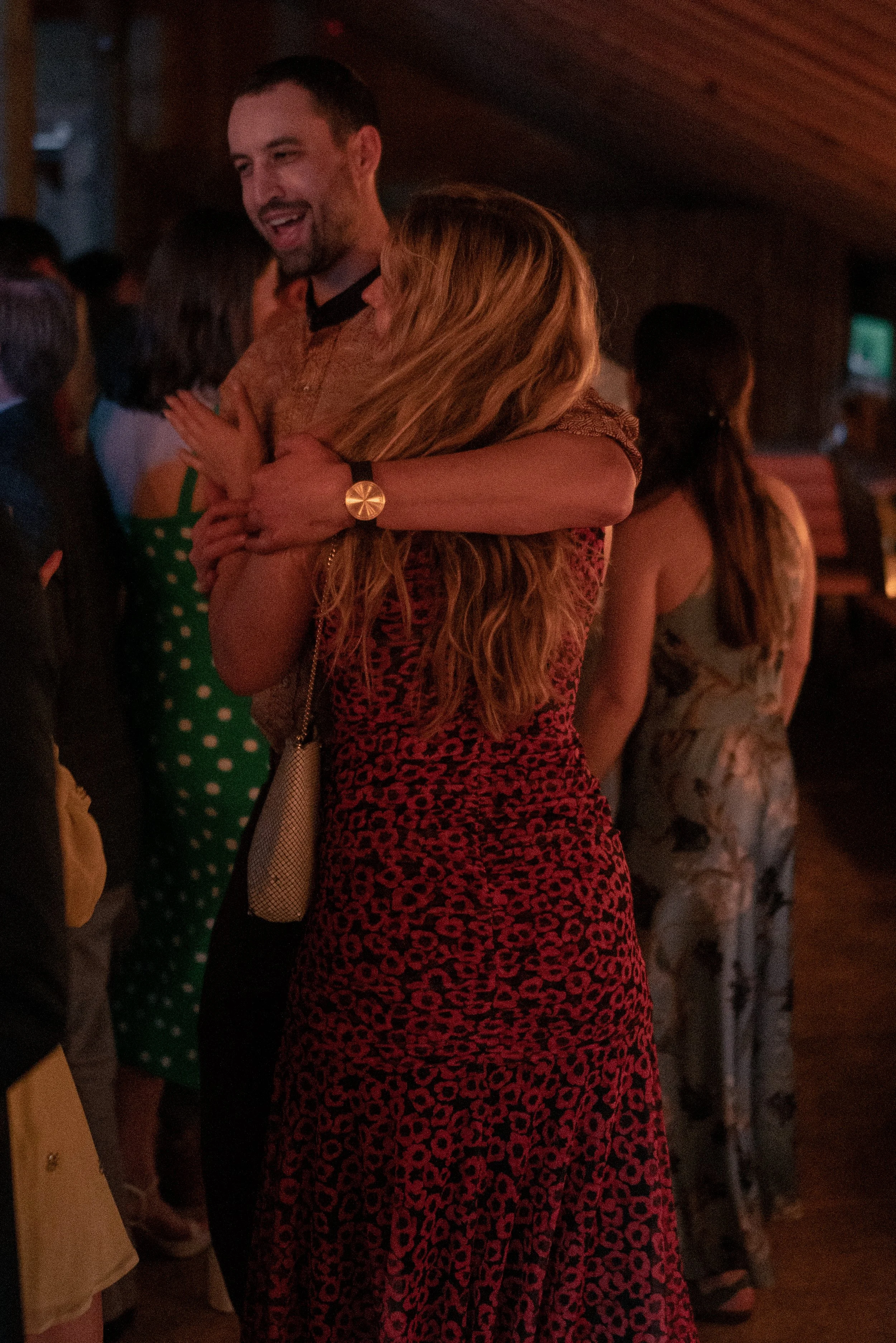 A man and a woman are hugging at a social gathering or party in a dimly lit room. The man is smiling with his eyes closed, and the woman has long, wavy hair. There are other people in the background, some wearing patterned dresses.