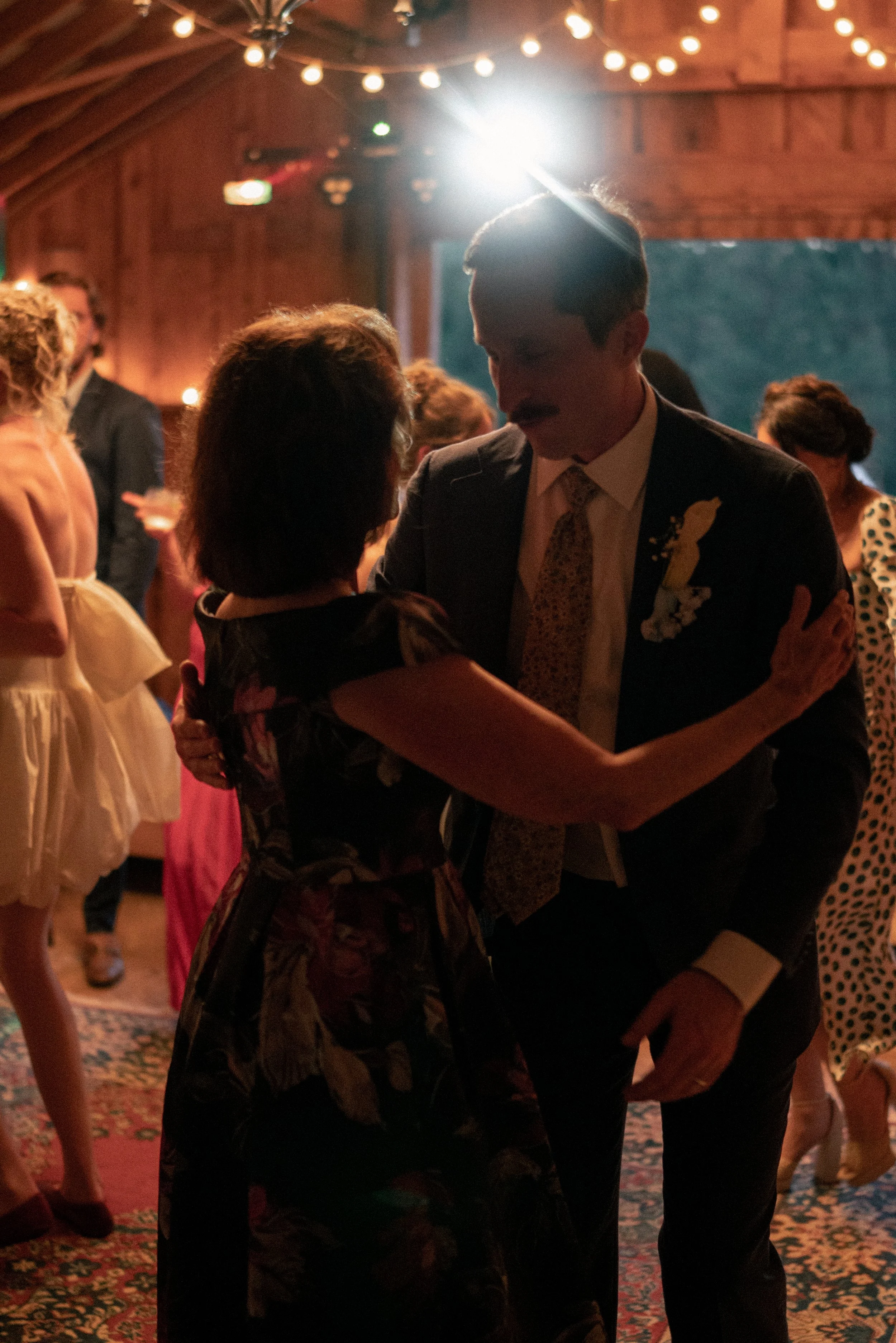 A man with a mustache in a suit and tie is dancing with a woman at a social event in a decorated venue with string lights. Other people are mingling in the background.