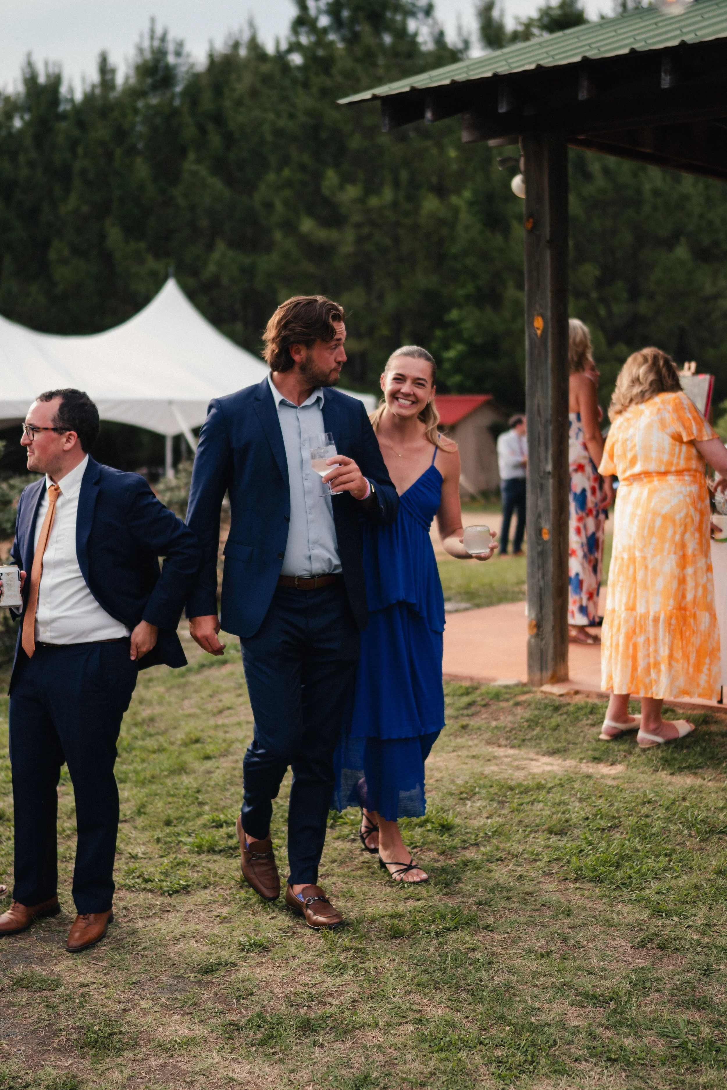 Group of people dressed in formal and semi-formal attire walking outdoors at a social gathering, with some holding drinks, in a grassy area near tents and a small building, during evening or late afternoon.