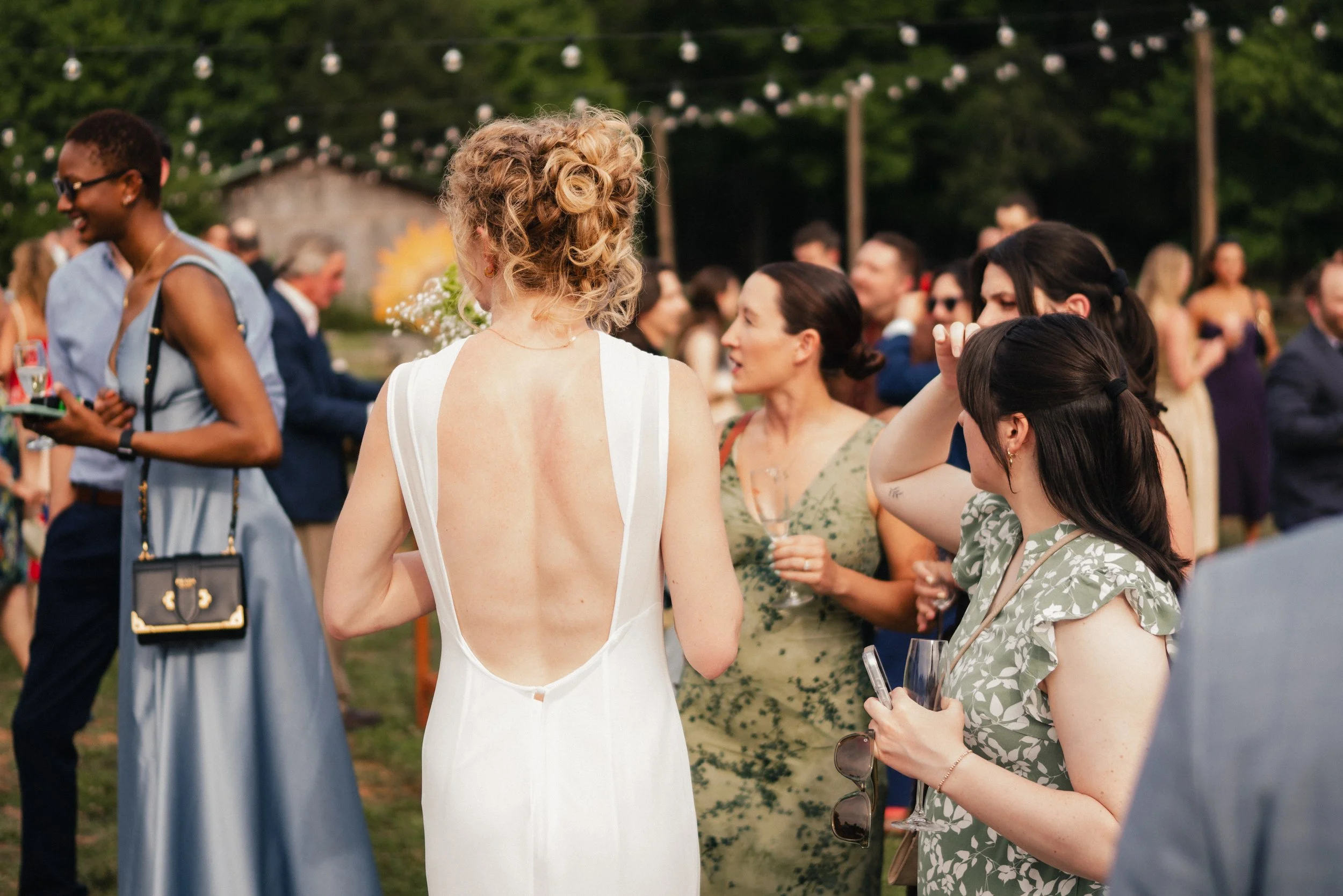 A group of women at an outdoor social event, including a woman with curly blonde hair in a white dress, talking to others holding wine glasses, with string lights and trees in the background at sunset.