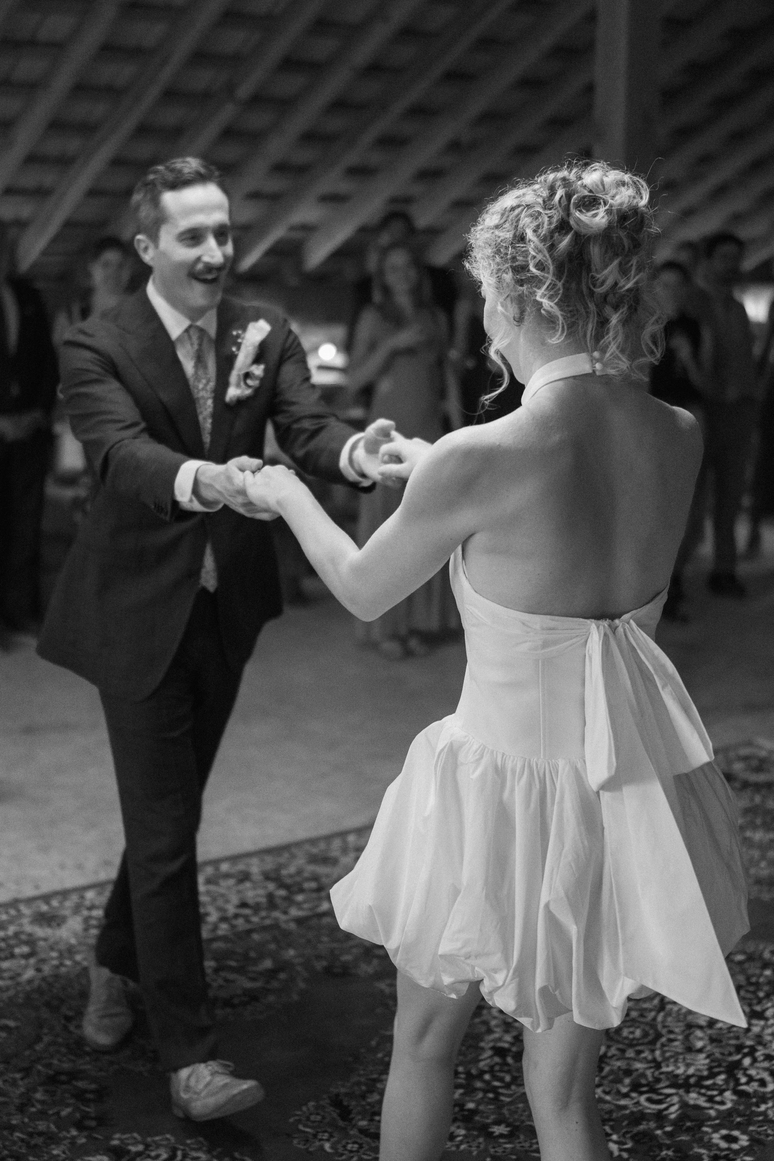 A joyful groom in a suit and a bride in a dress dancing during their wedding reception inside a wooden venue.