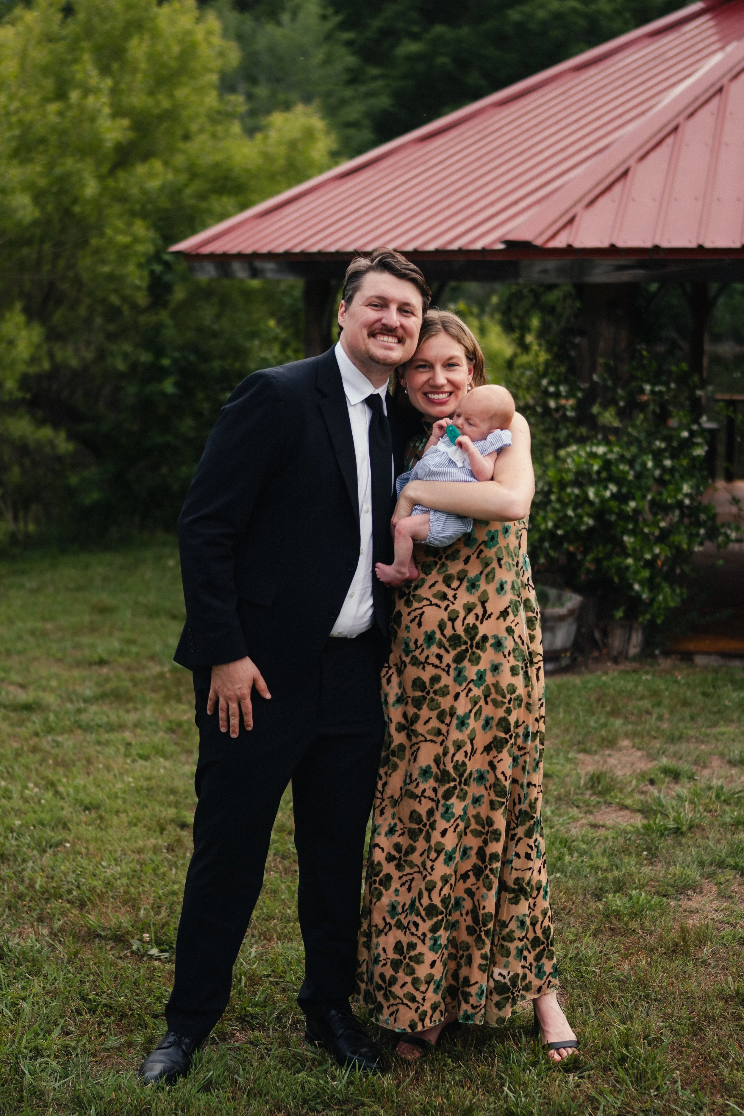 A smiling couple dressed in formal attire stands outdoors on grass near a wooden structure with a red roof, holding a baby dressed in light blue and white striped clothing.