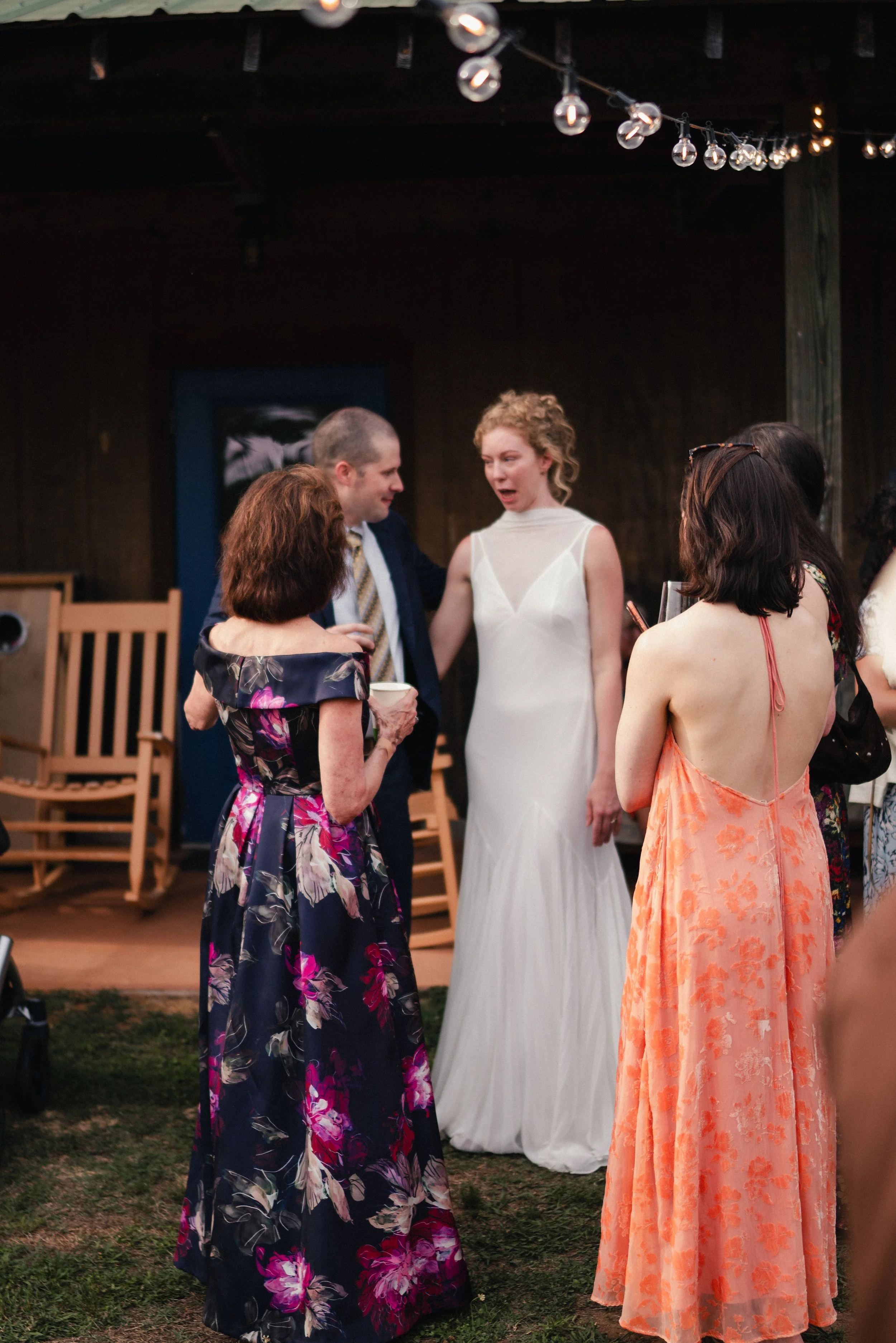 People gathered outdoors at a wedding reception, conversing and celebrating under string lights, with a woman in a white wedding dress and a man in a dark suit and striped tie in the background.