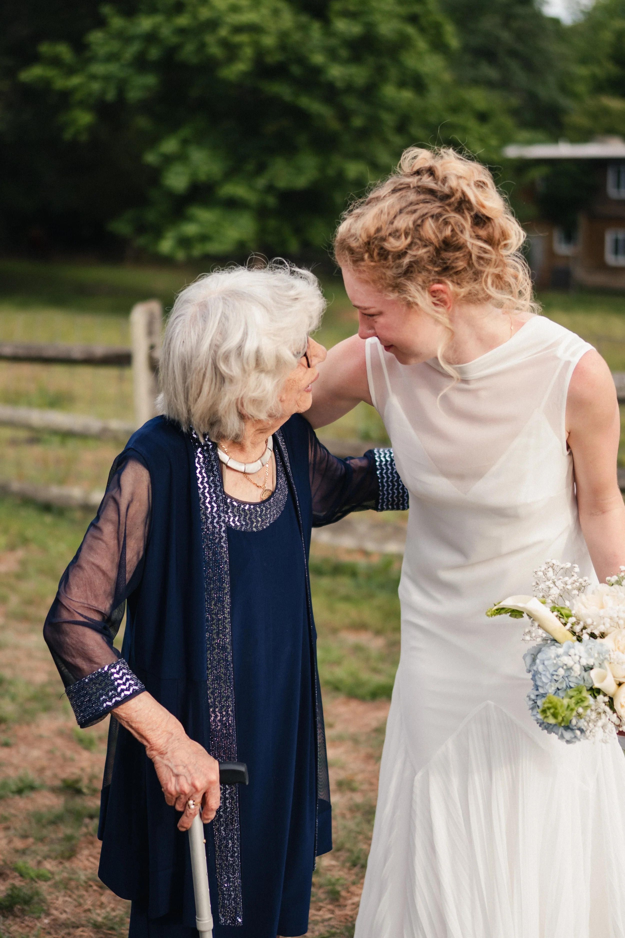 A young woman in a white wedding dress leaning toward an elderly woman in a navy dress with intricate beading, sharing an emotional moment outdoors near a wooden fence with greenery in the background.