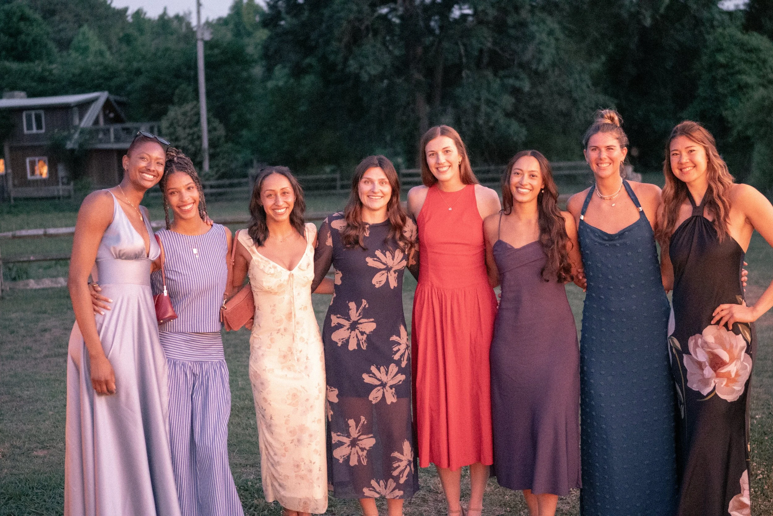 Group of eight women standing outdoors on grass, smiling, with trees and a house in the background during dusk.