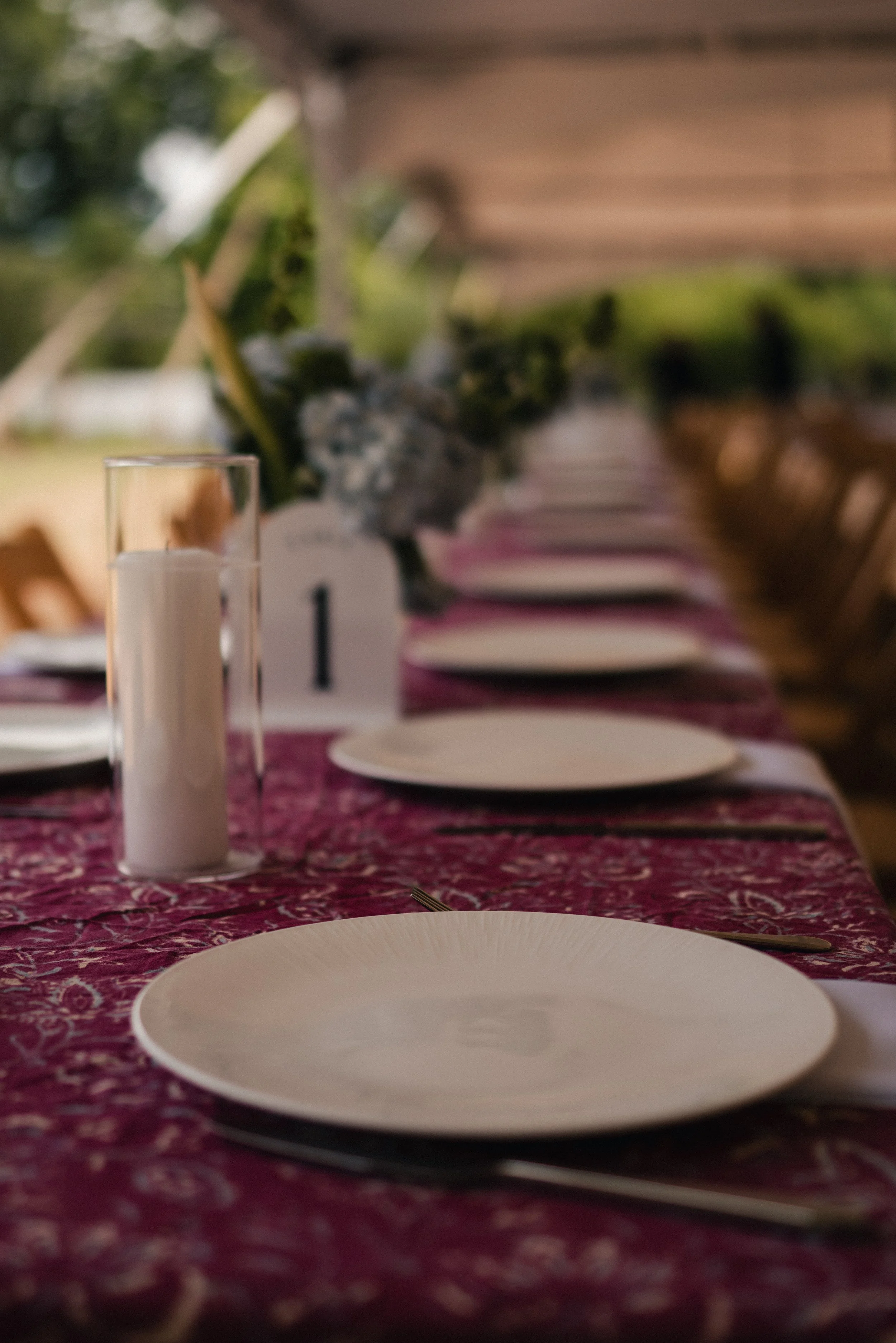 A long dining table set for a meal with white plates, silverware, and a purple patterned tablecloth. There are glass candle holders with white candles and a floral centerpiece in the background.