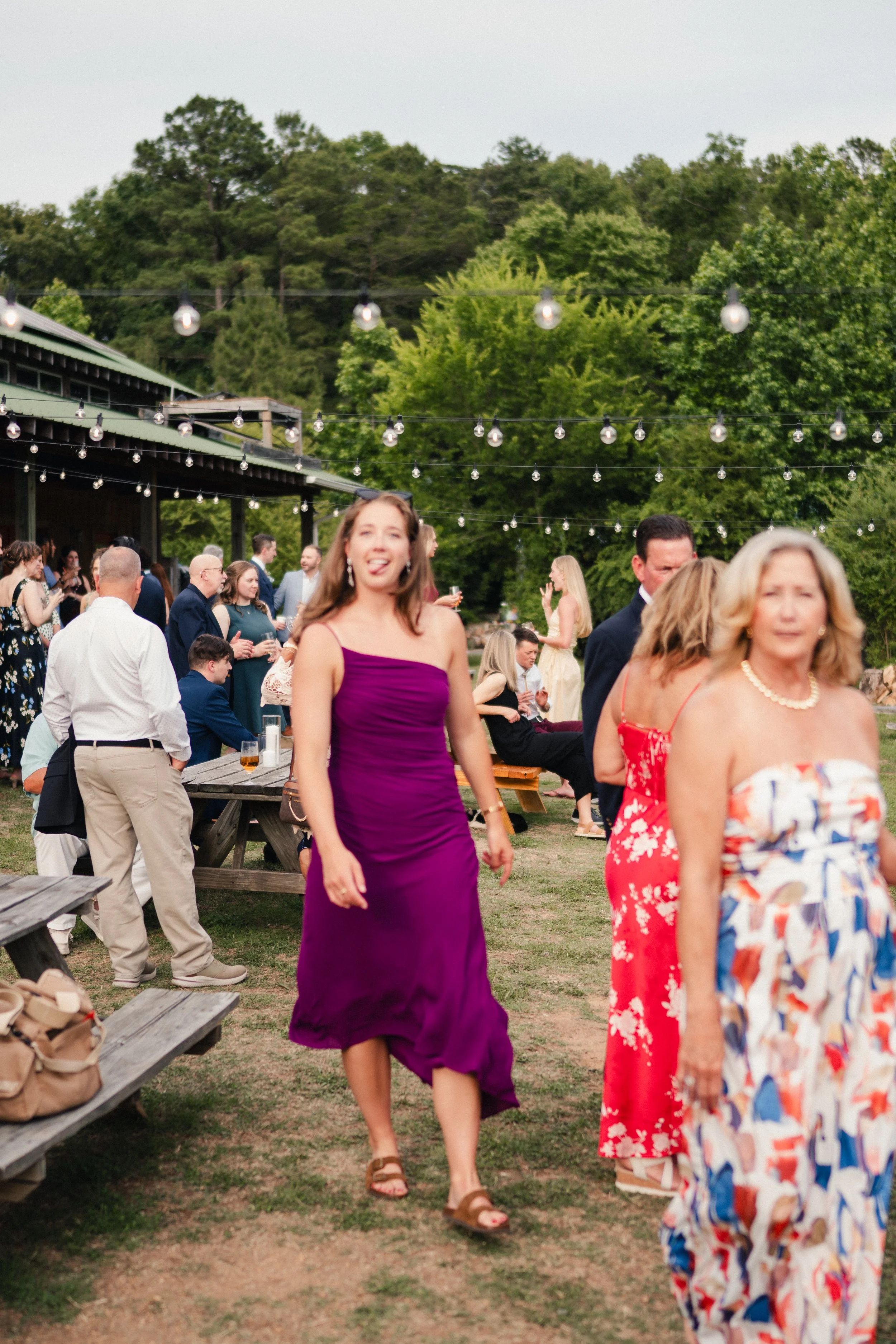 People at an outdoor gathering or celebration, with string lights hanging above, trees and a building in the background, and guests mingling and walking.