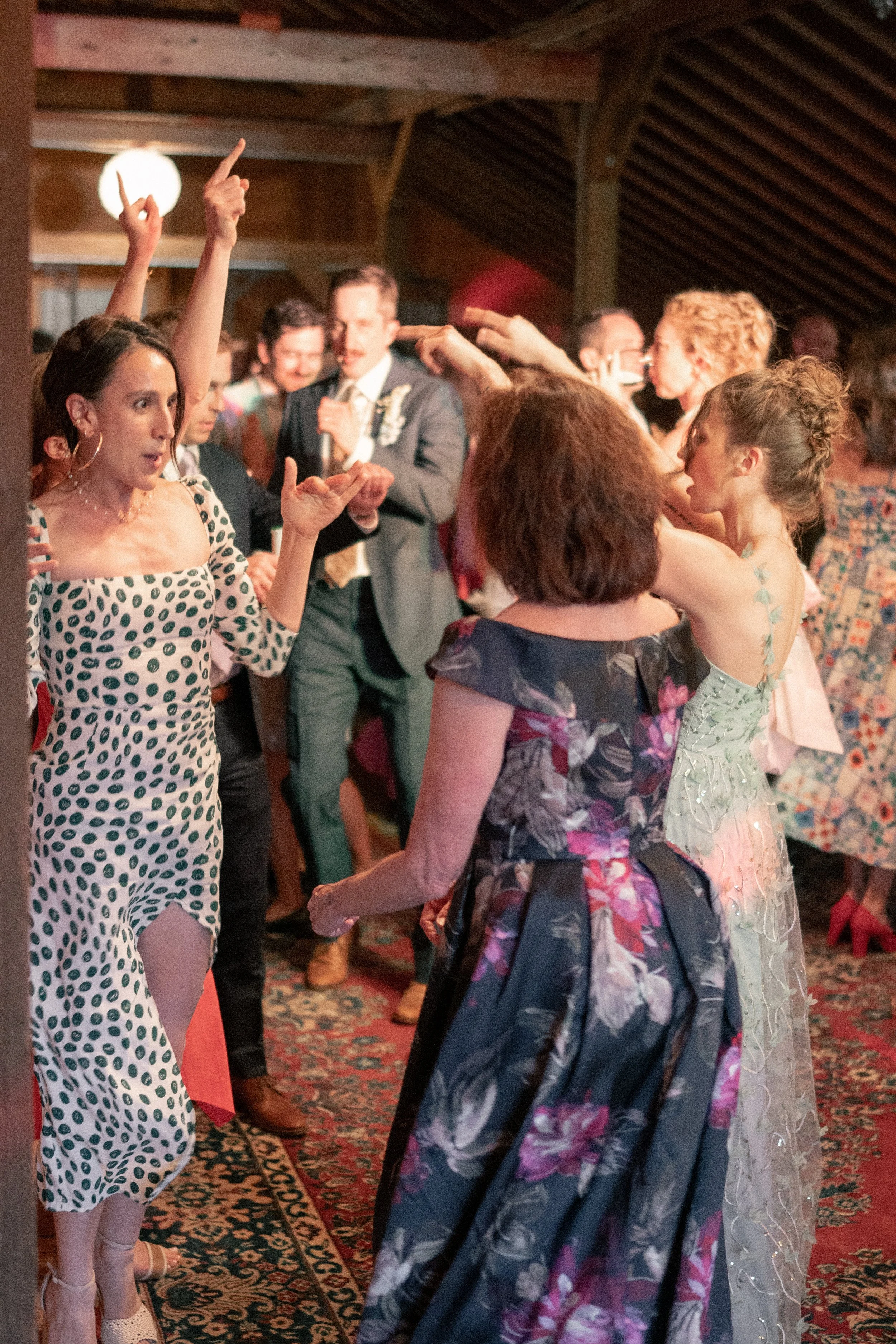 A group of people dancing at a lively party. A woman in a polka dot dress is in the foreground, gesturing with her hands, while others in the background are smiling and talking.