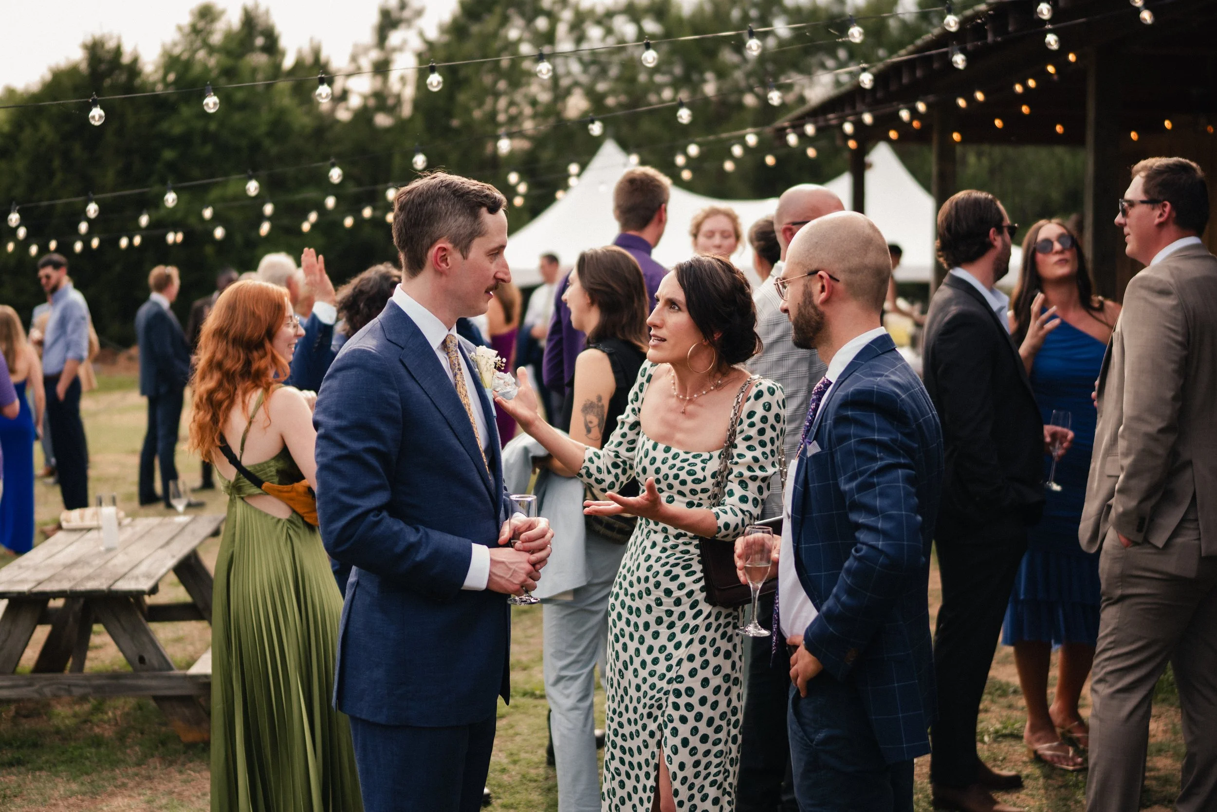 People at an outdoor social event, talking and mingling under string lights in the evening.