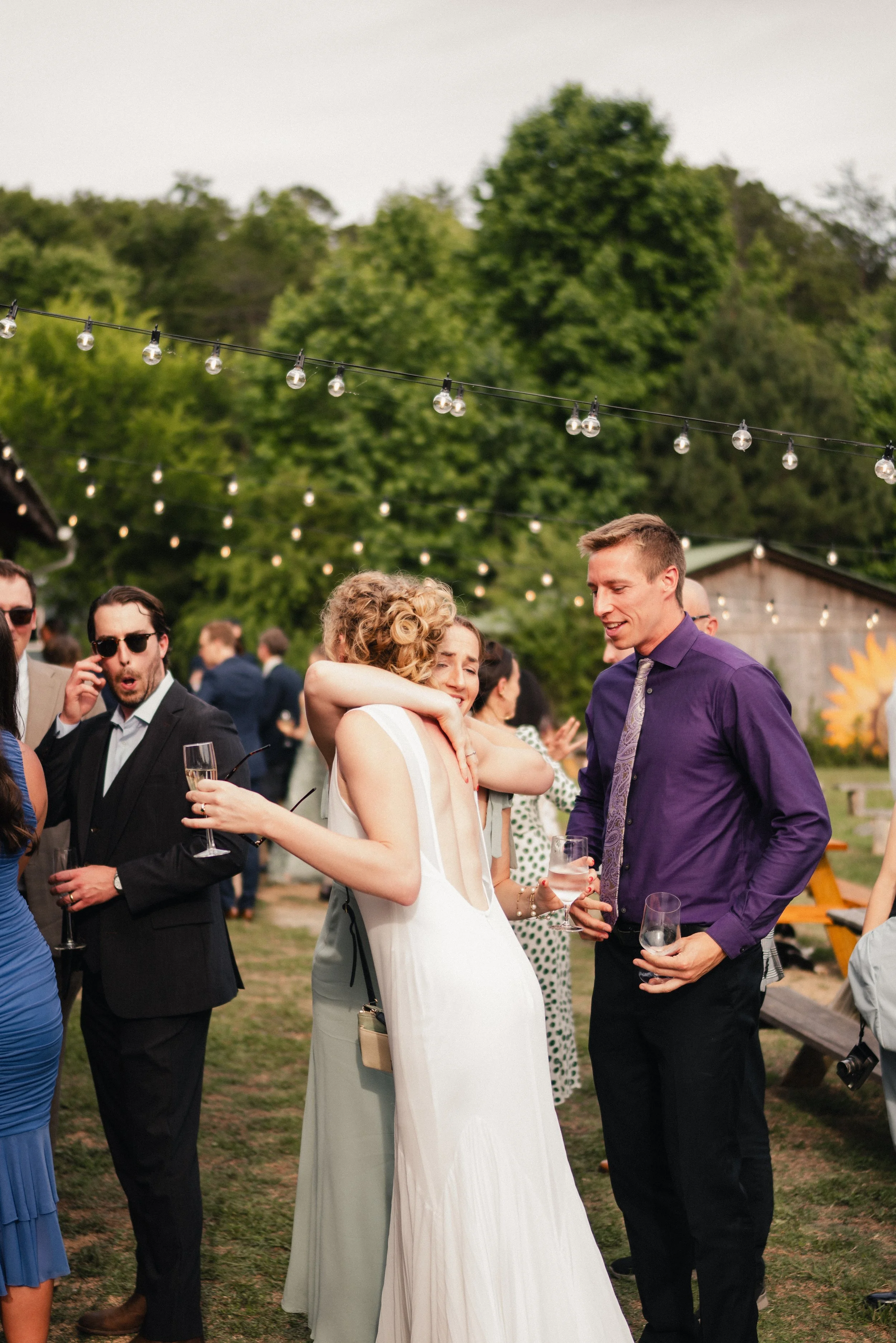 People celebrating outdoors at an event resembling a wedding reception, with string lights overhead and trees in the background.