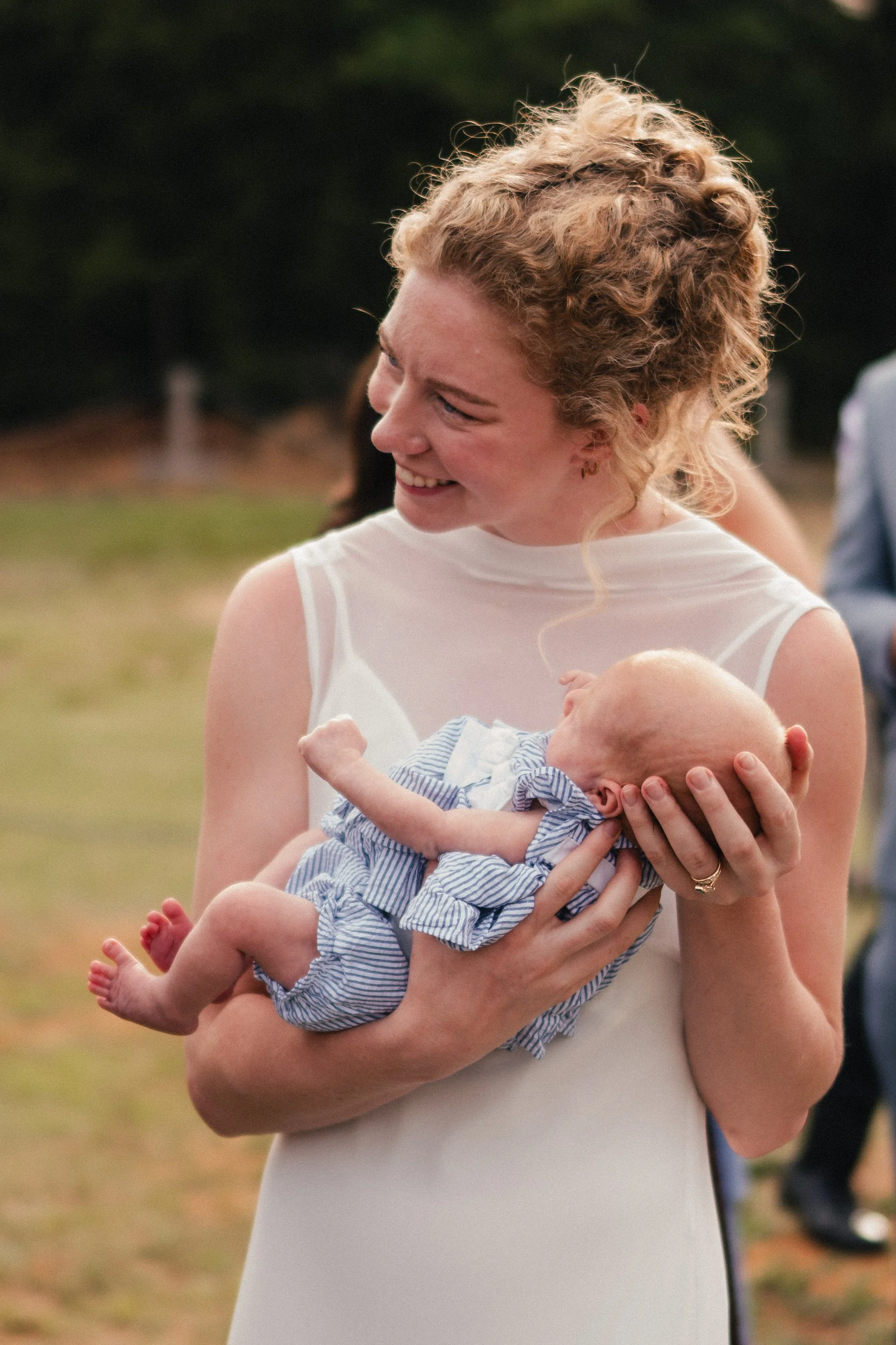 A woman with curly blonde hair smiling and holding a baby in a striped outfit outdoors.