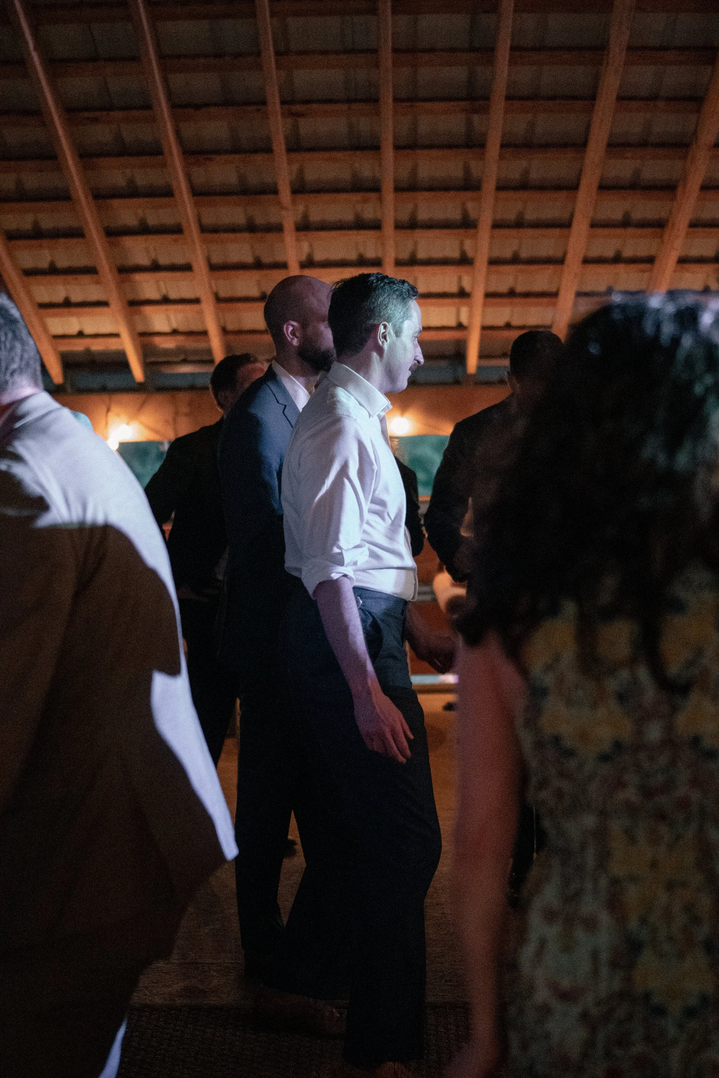Group of people standing together in an indoor setting with a wooden roof structure, some individuals are dressed in formal attire, engaged in conversation or listening.