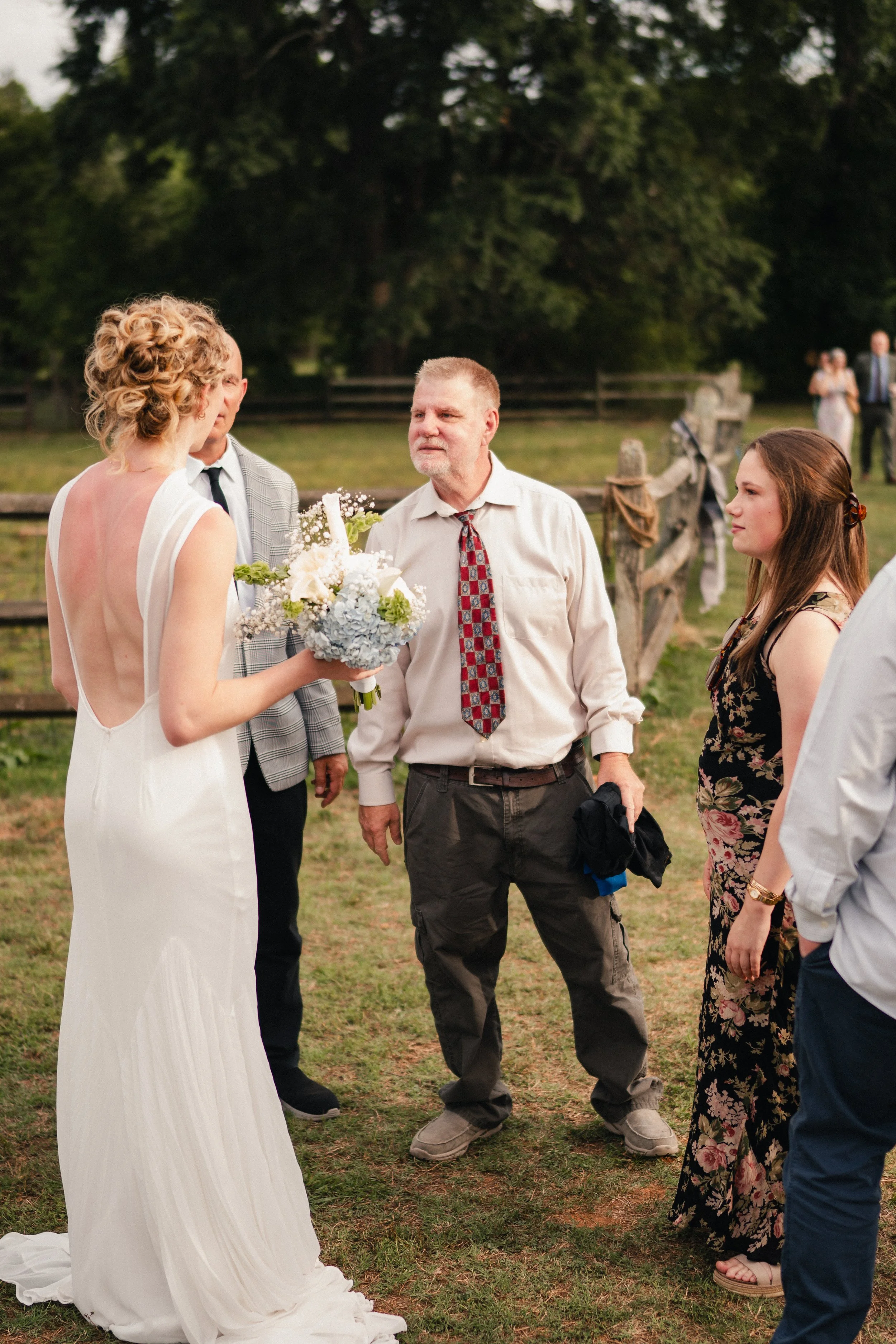 A wedding scene outdoors with a bride holding a bouquet talking to an older man, surrounded by several guests, with a wooden fence and trees in the background.
