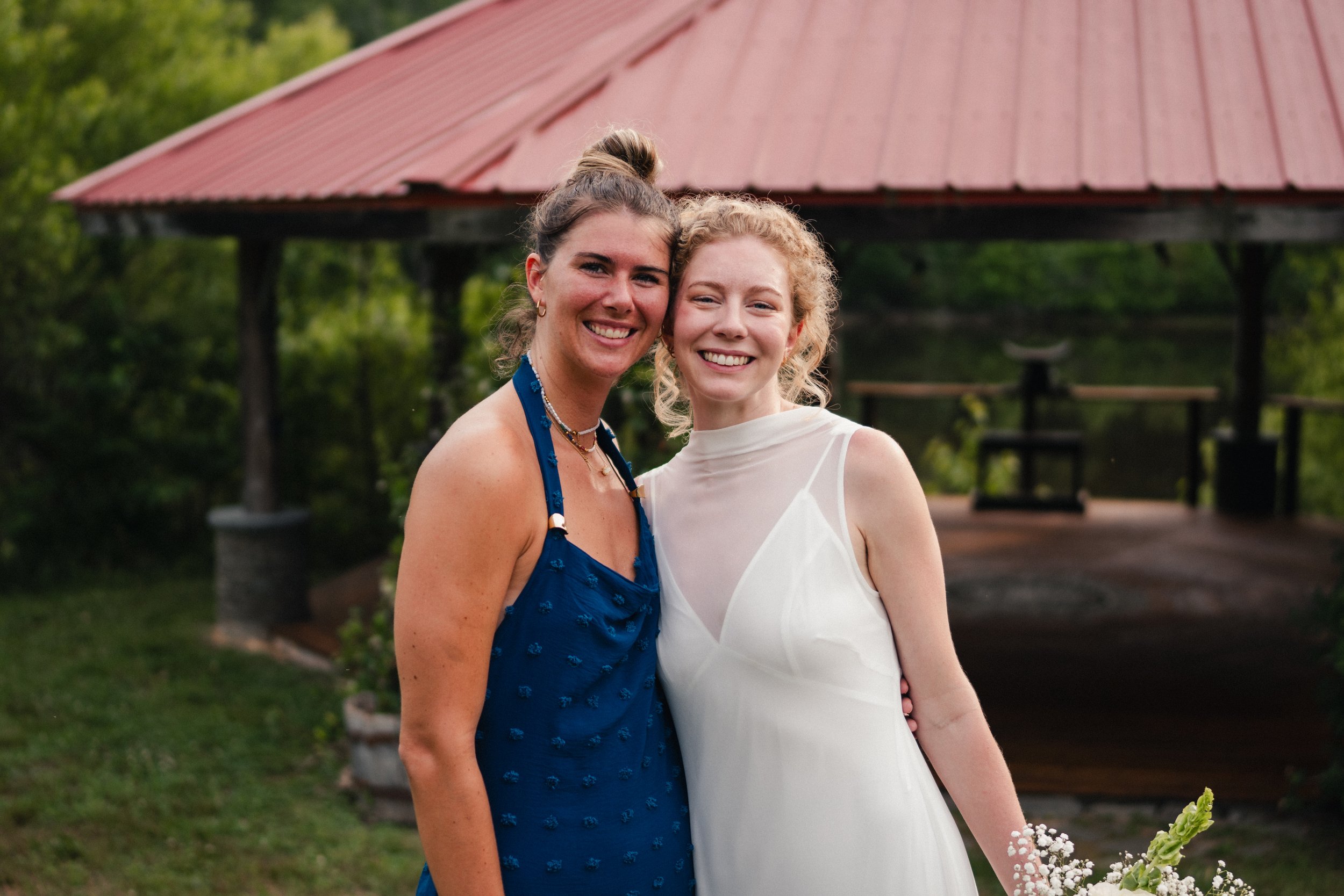 Two women smiling and embracing each other outdoors in front of a red-roofed pavilion, with greenery in the background.