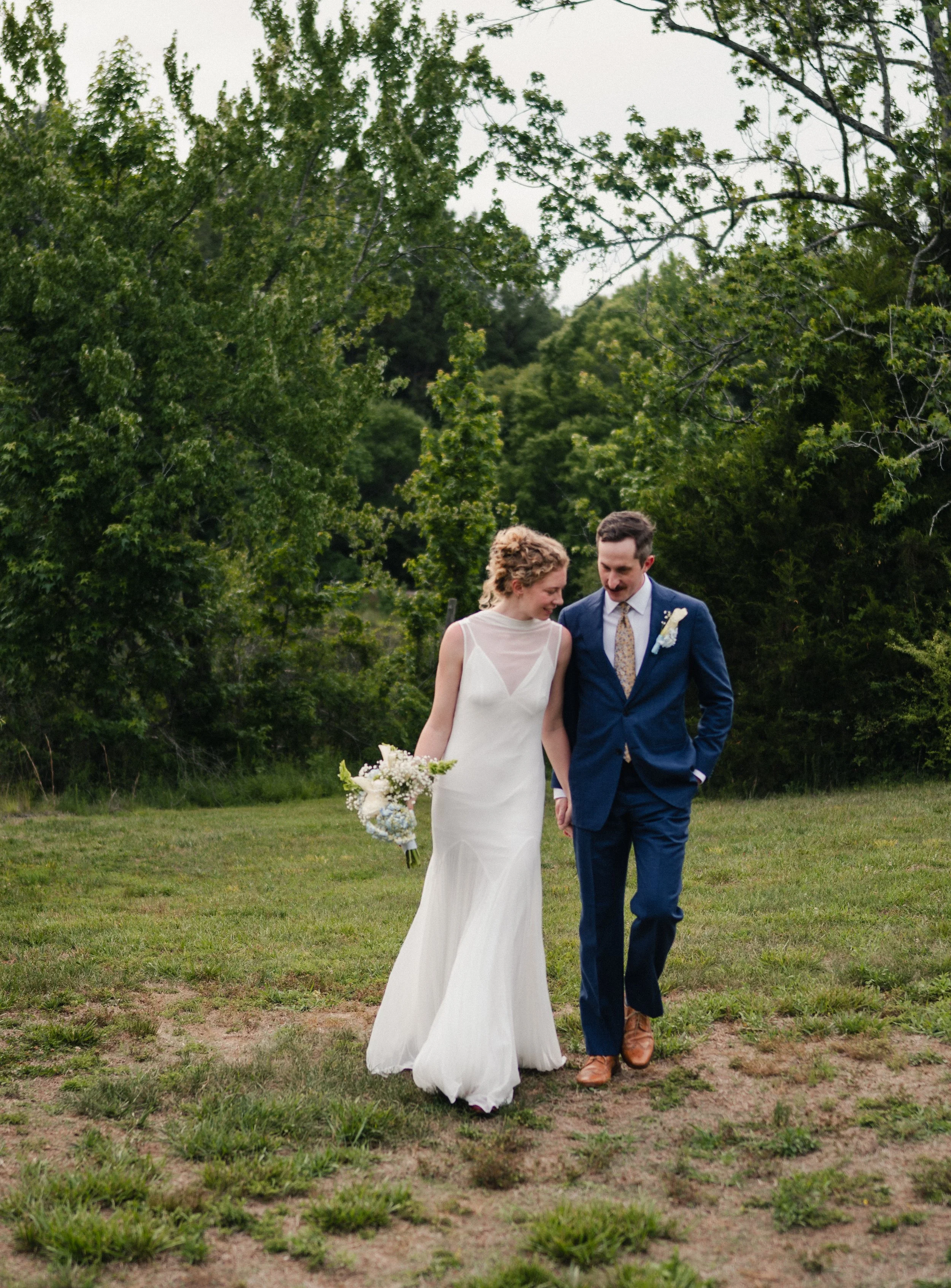 A bride and groom walking together outdoors on a grassy field, holding hands, with trees in the background. The bride is wearing a white wedding dress holding a bouquet, and the groom is wearing a navy suit with a boutonnière.
