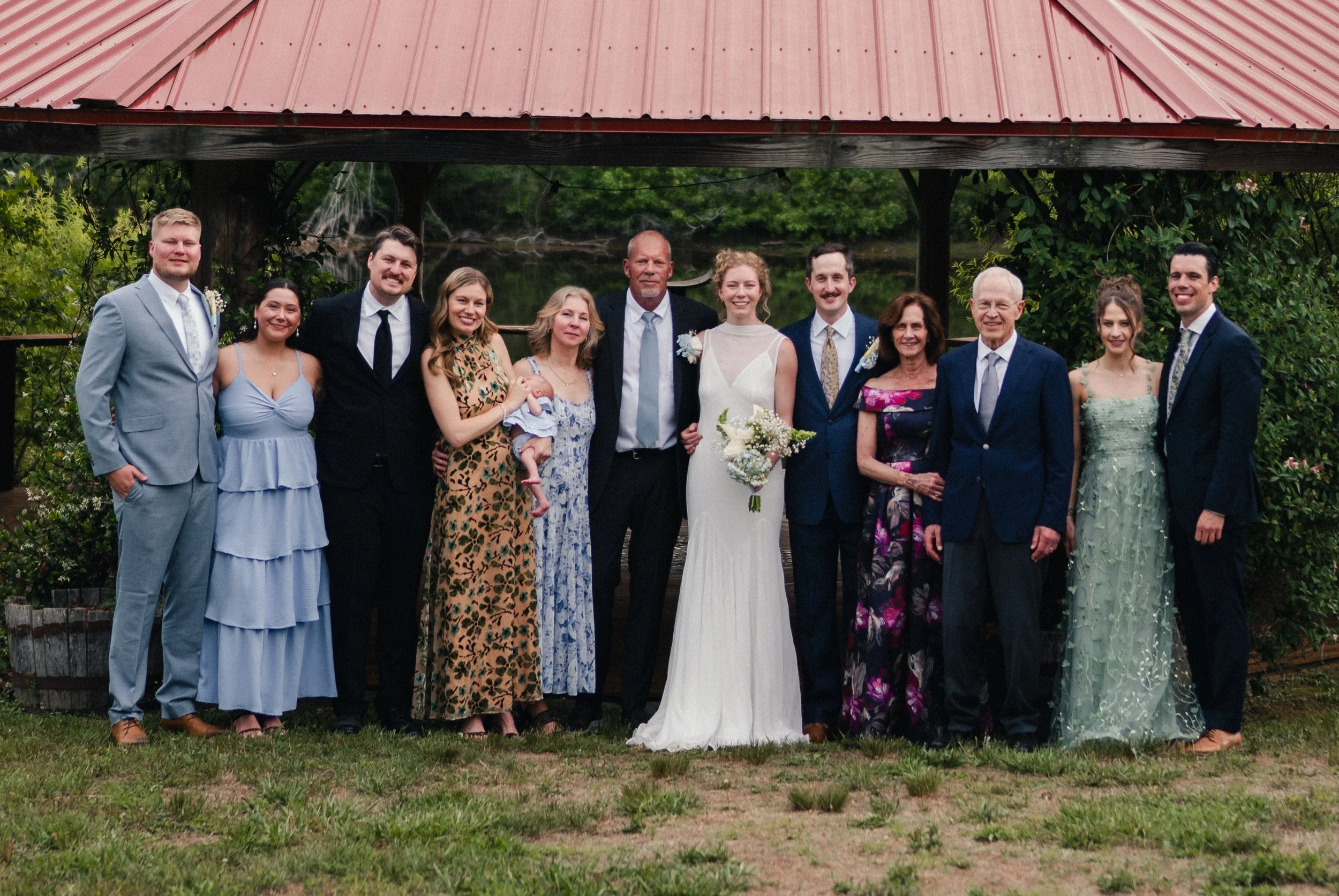 A group of people dressed in formal and semi-formal attire standing outdoors, with a red roofed pavilion in the background, during a daytime celebration.