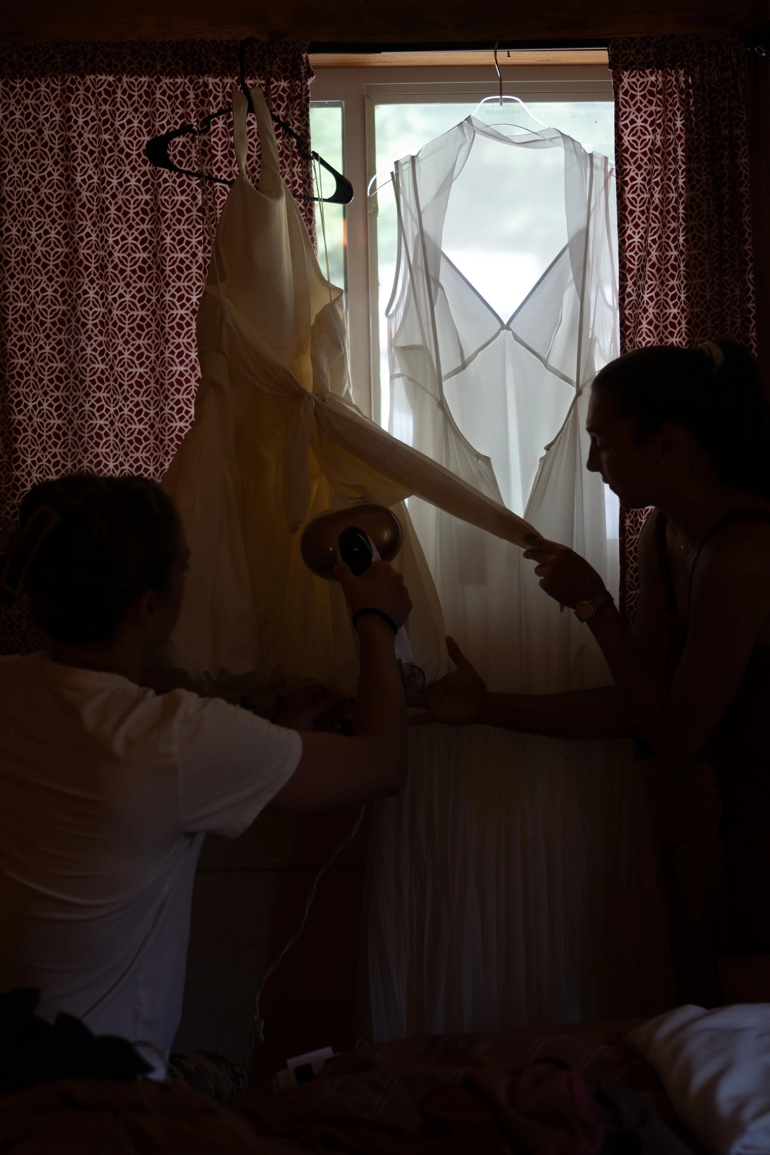 A window with pattern curtains has wedding dresses hanging in front of it, one with a sheer fabric and the other with a satin finish. Two people are in the foreground, one holding the train of a wedding dress.