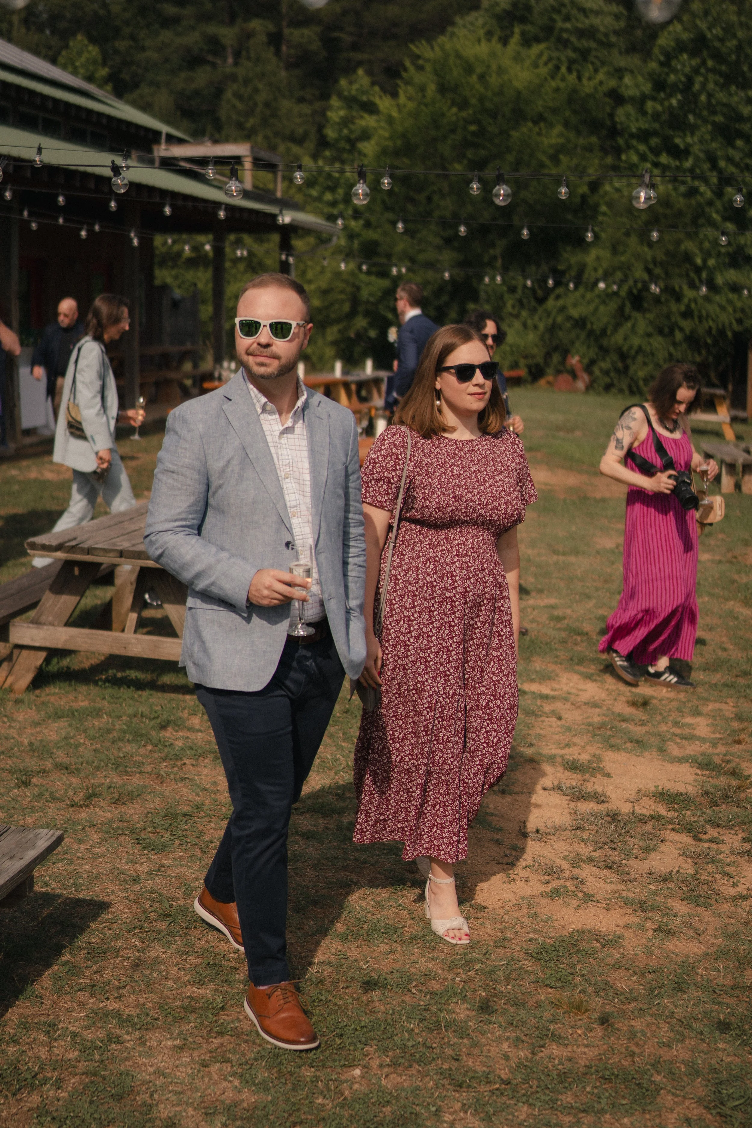 People at an outdoor gathering, two individuals in the foreground wearing sunglasses, a man in a light gray blazer and white shirt holding a drink, and a woman in a red patterned dress. Other guests are visible in the background near a wooden buildin