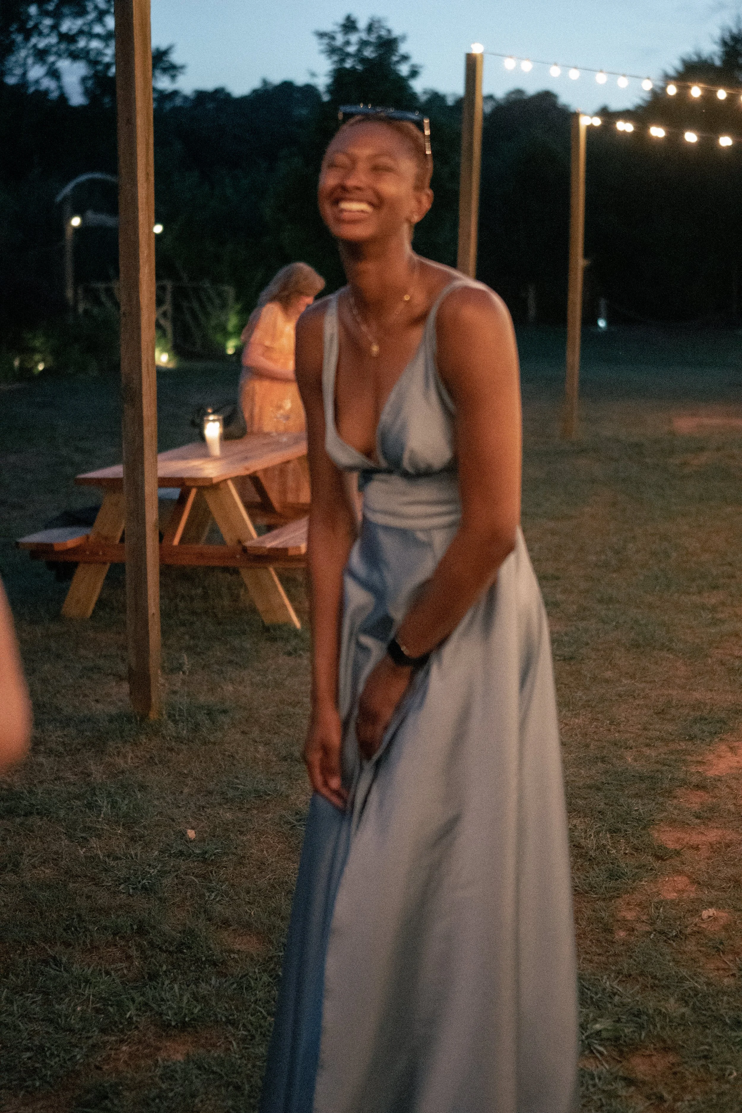 A woman in a light-colored dress smiling and looking to the side outdoors during evening with string lights hanging above and blurred background of trees and a person in a patterned dress.