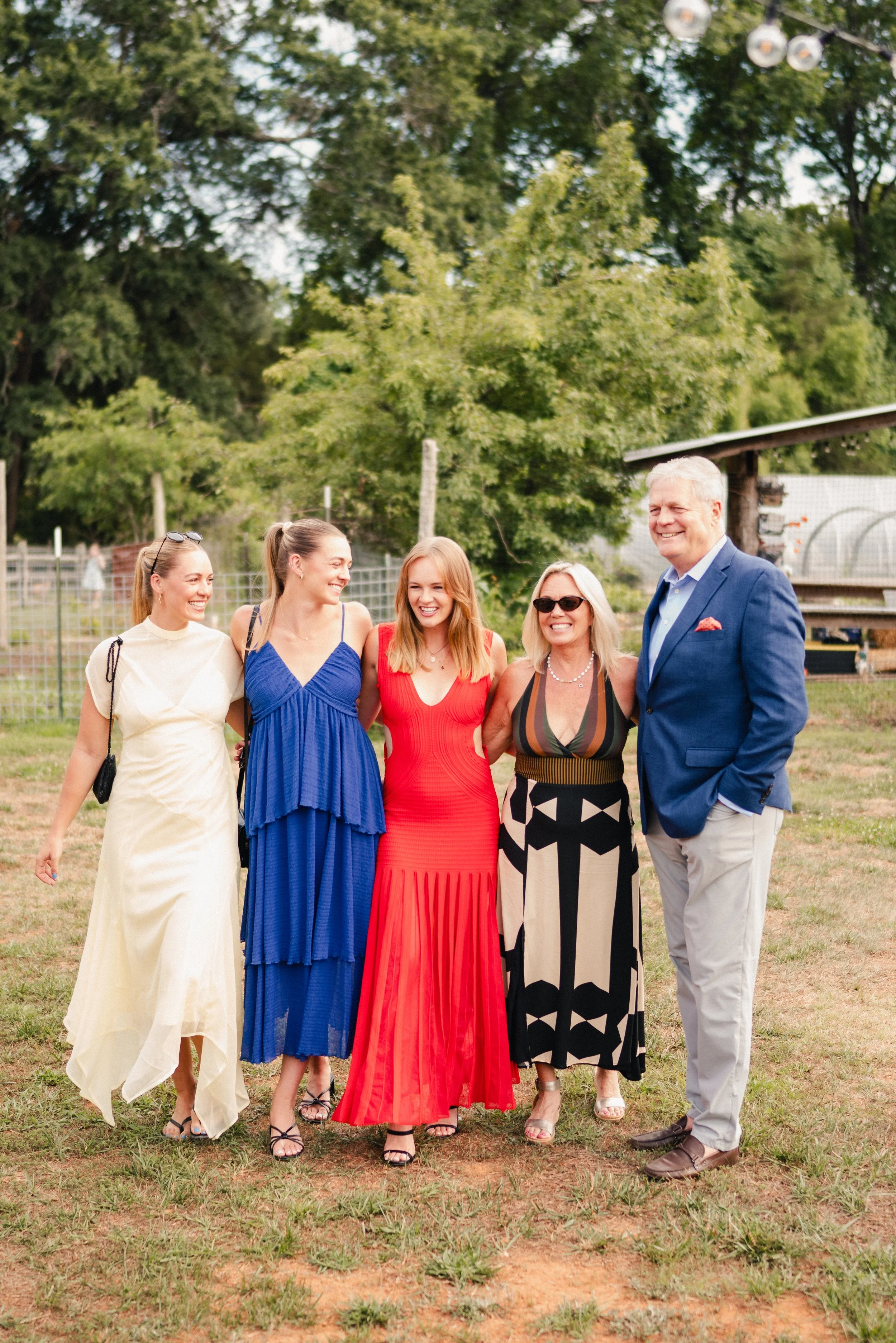 A group of five women and one man standing outdoors on grass, smiling and posing for a photo. The women are dressed in colorful summer dresses, and the man is in a blue suit. There are trees and a fence in the background.
