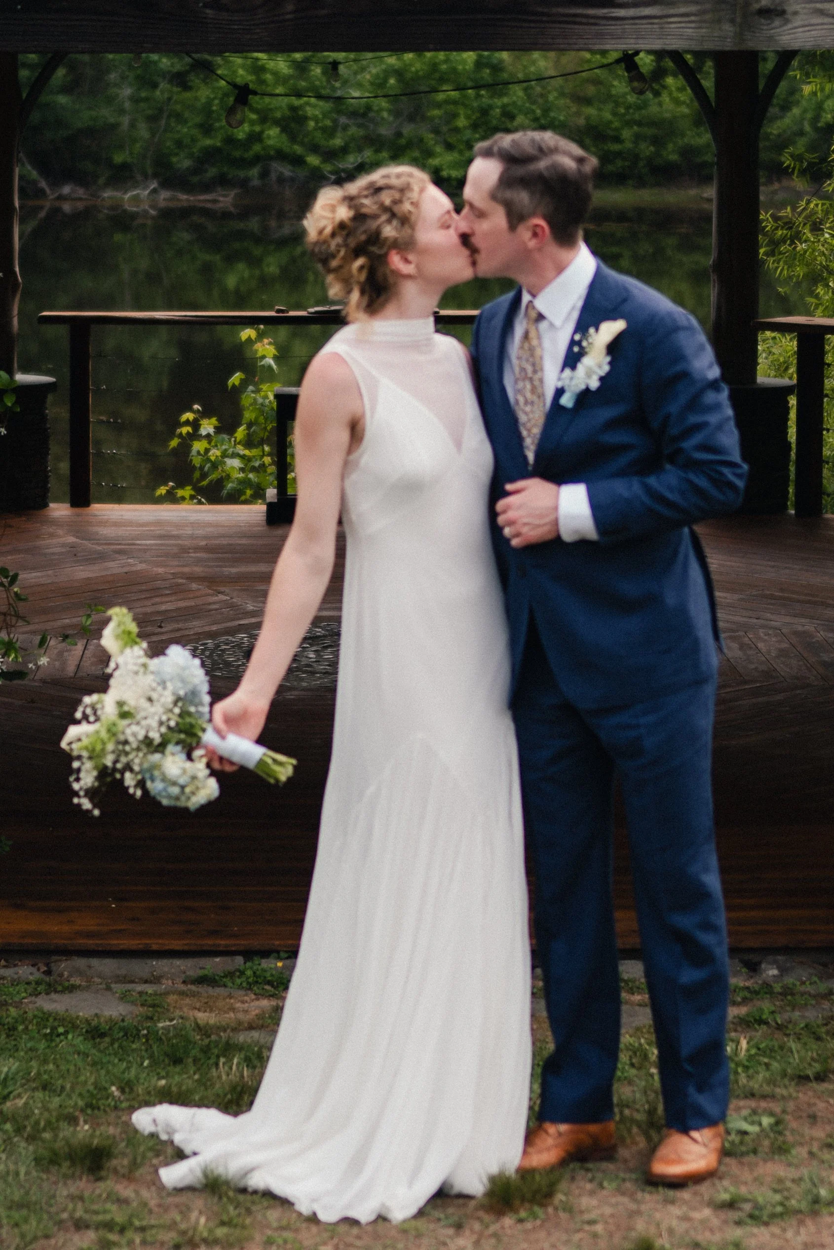 A newlywed couple sharing a kiss outdoors on a wooden deck, the bride in a white dress holding a bouquet of flowers, and the groom in a blue suit with a boutonniere, surrounded by greenery and string lights overhead.