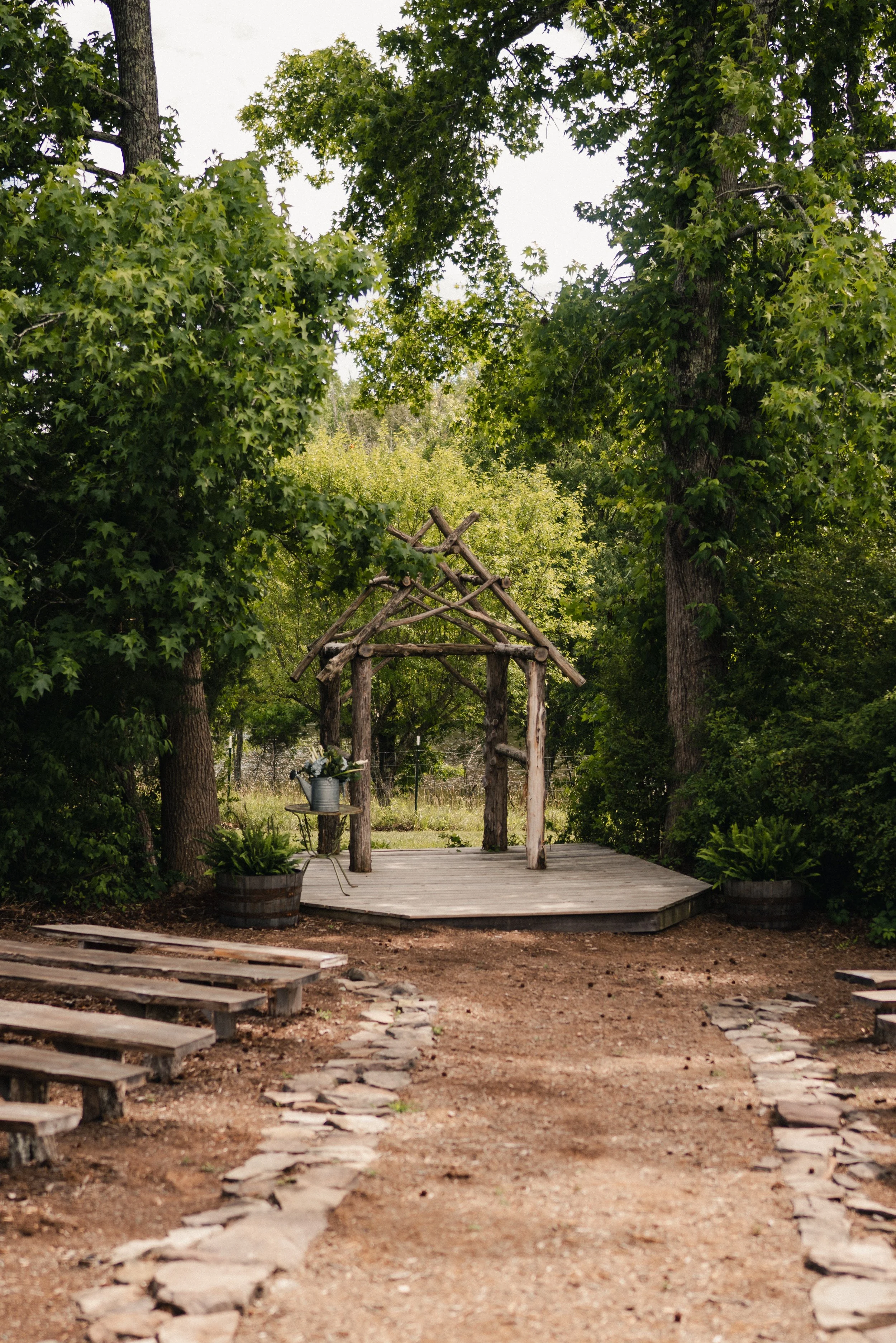 A small wooden wedding arch on a wooden platform, surrounded by lush green trees, with stone pathway and planters in the foreground.