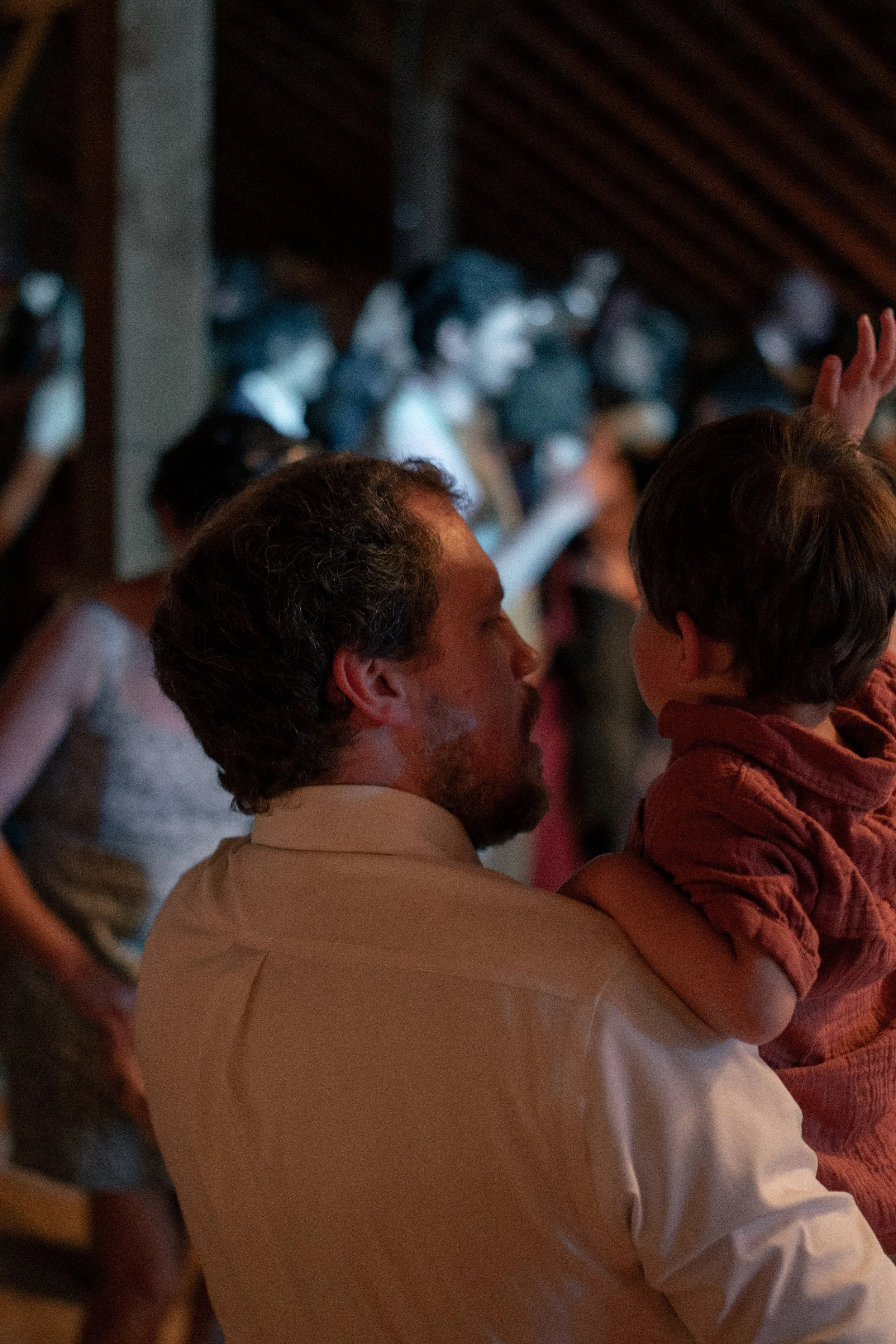 A man holding a young boy in a crowded room with dim lighting, engaged in conversation.