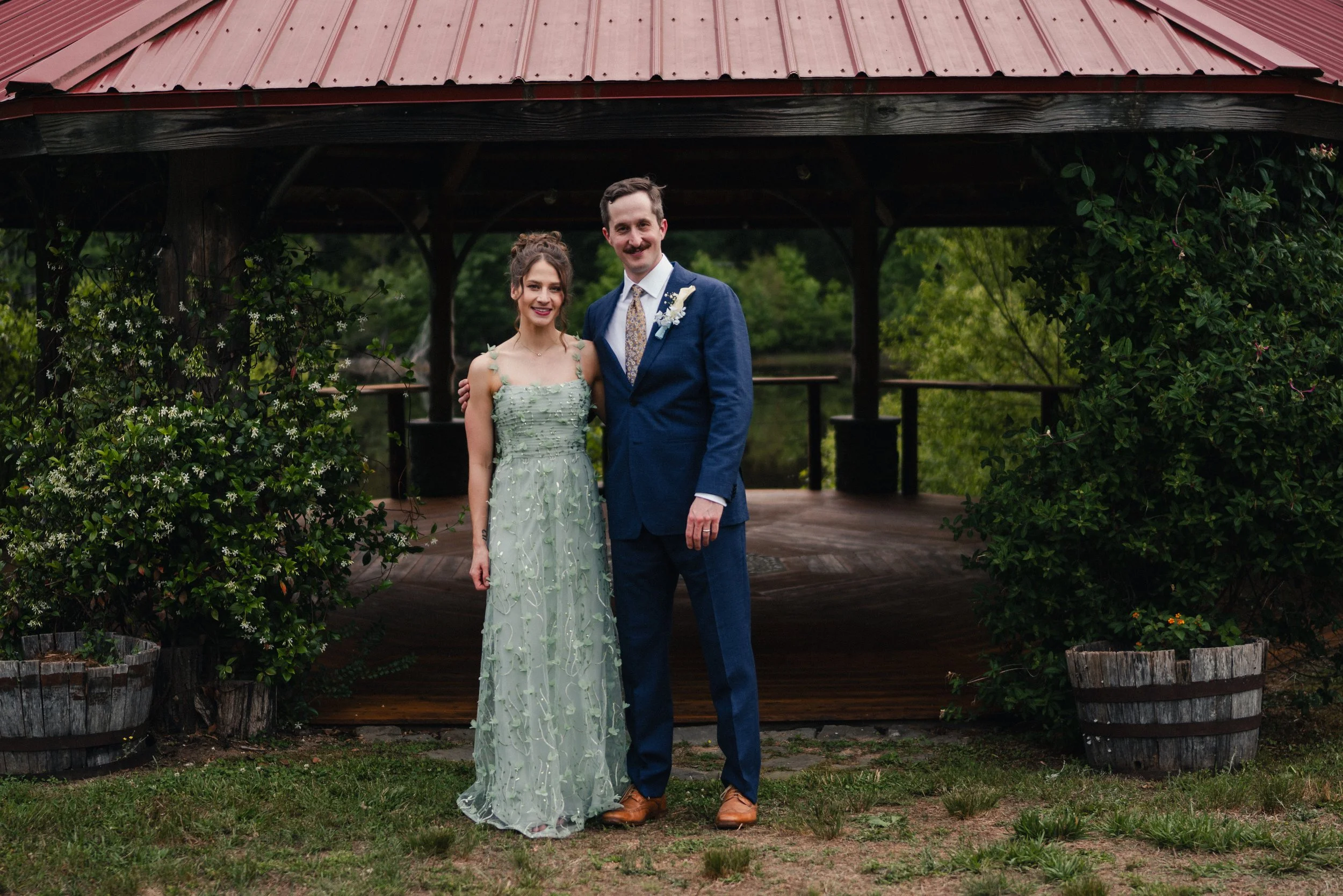 A couple dressed in formal attire standing outdoors under a wooden pavilion with a red metal roof, surrounded by greenery and shrubs.