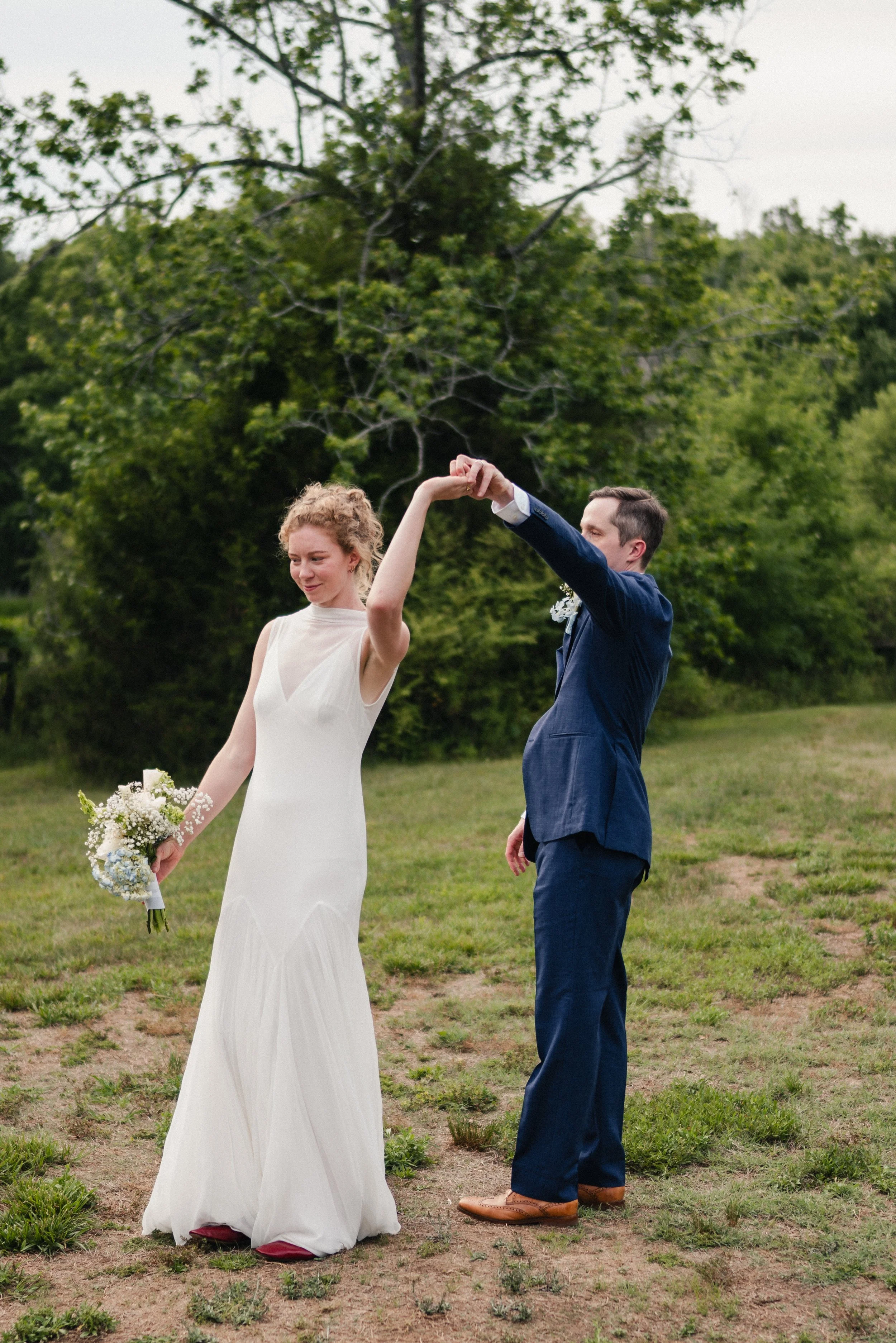 A bride and groom holding hands and dancing outdoors on a grassy field with green trees in the background.