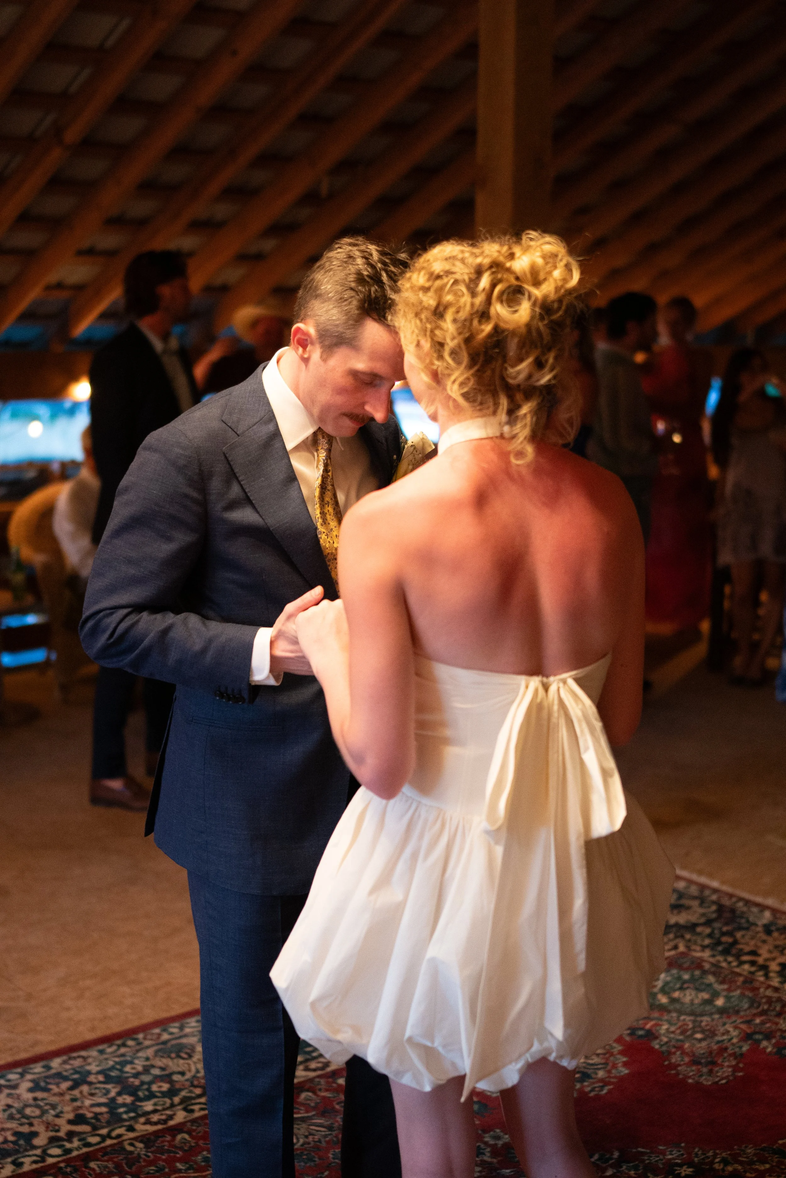 A man and a woman dancing closely at an indoor event. The man is wearing a dark suit with a patterned tie, and the woman is wearing a strapless cream-colored dress with a bow at the back. The background shows other people socializing and a wooden, va