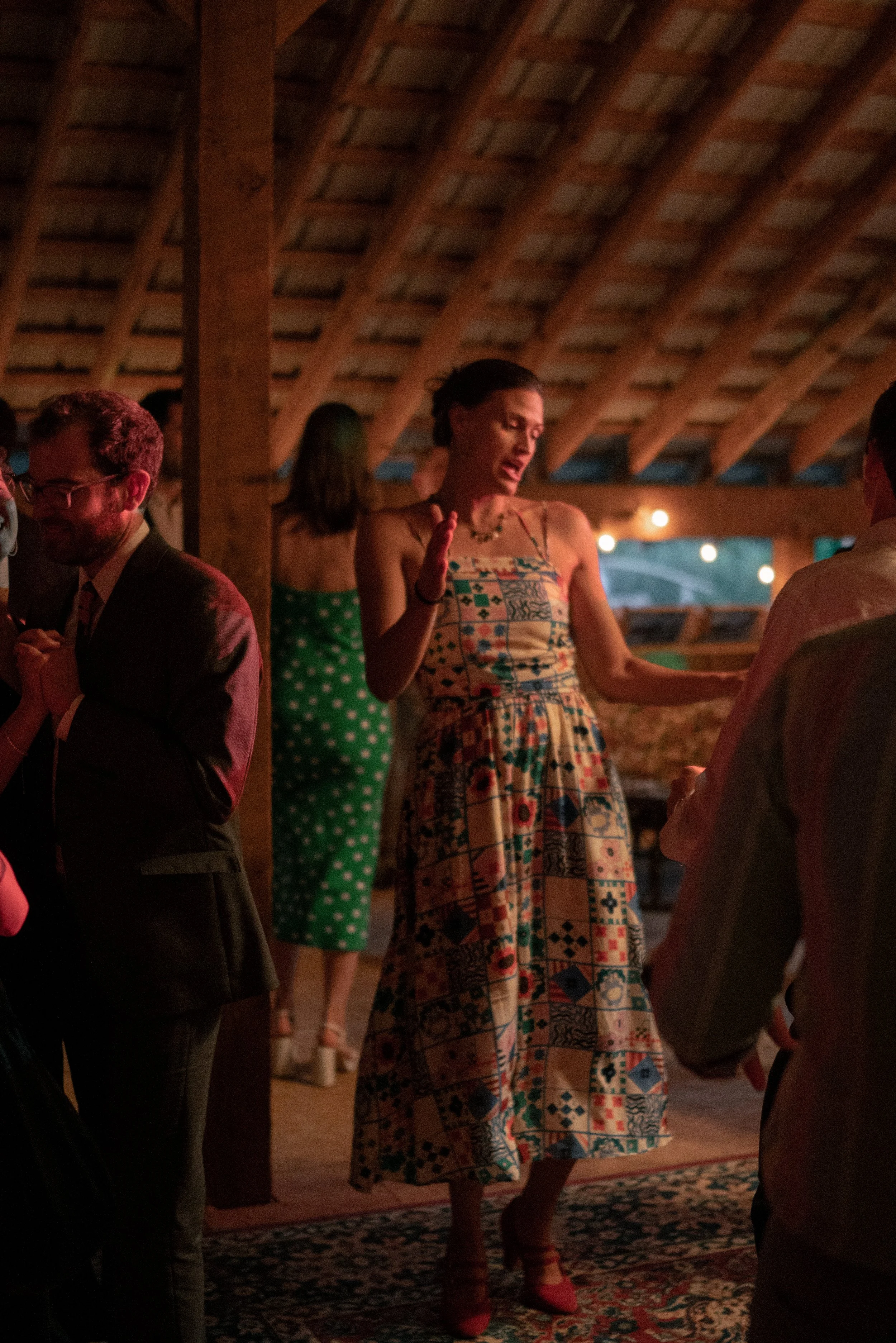 A woman in a colorful patterned dress and red heels appears to be singing or speaking passionately at a gathering in a dimly lit, rustic wooden venue. Several other people are present, some with phones or engaged in conversations.