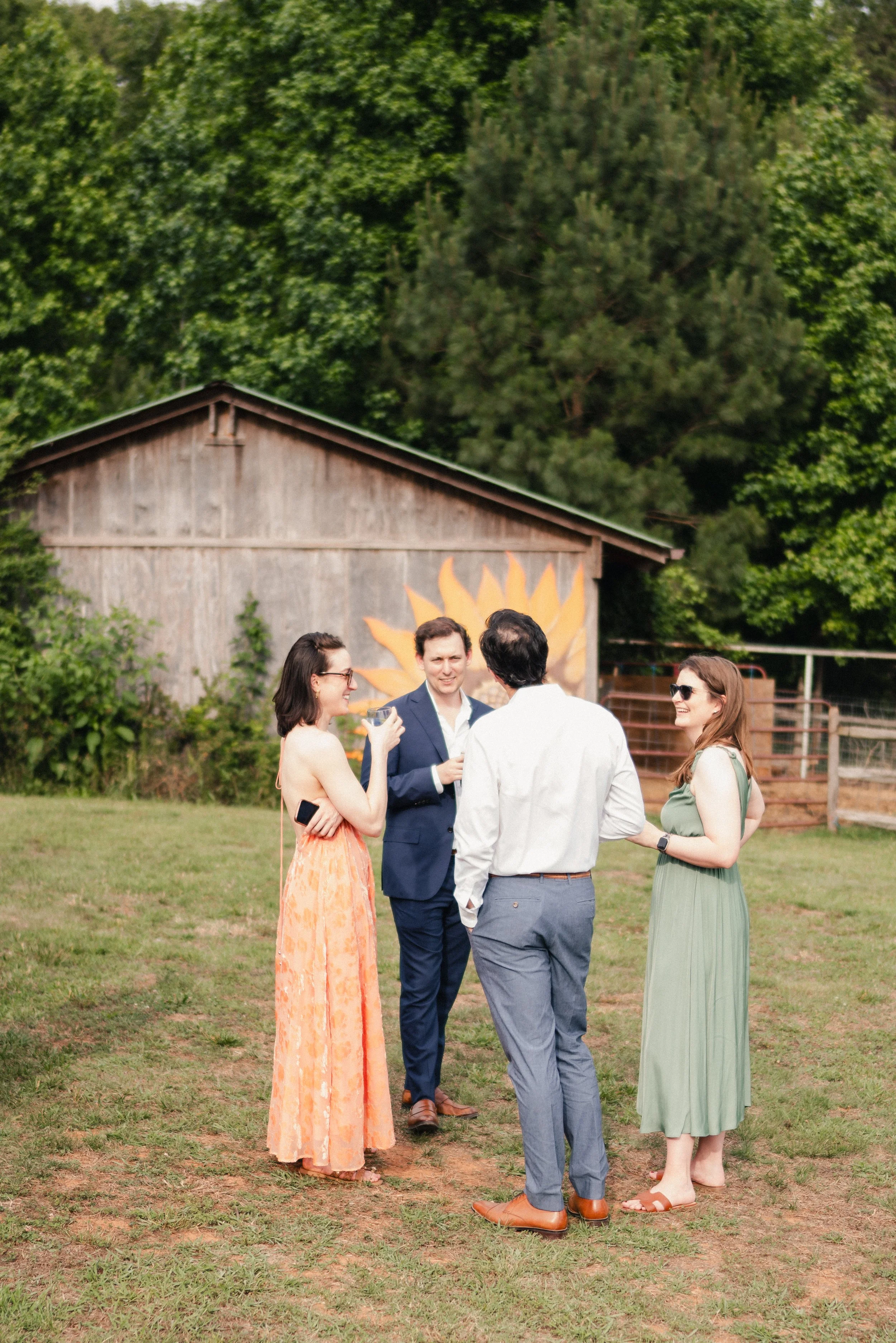 Group of five people standing on grass at an outdoor event, engaged in conversation, with a rustic barn and green trees in the background.