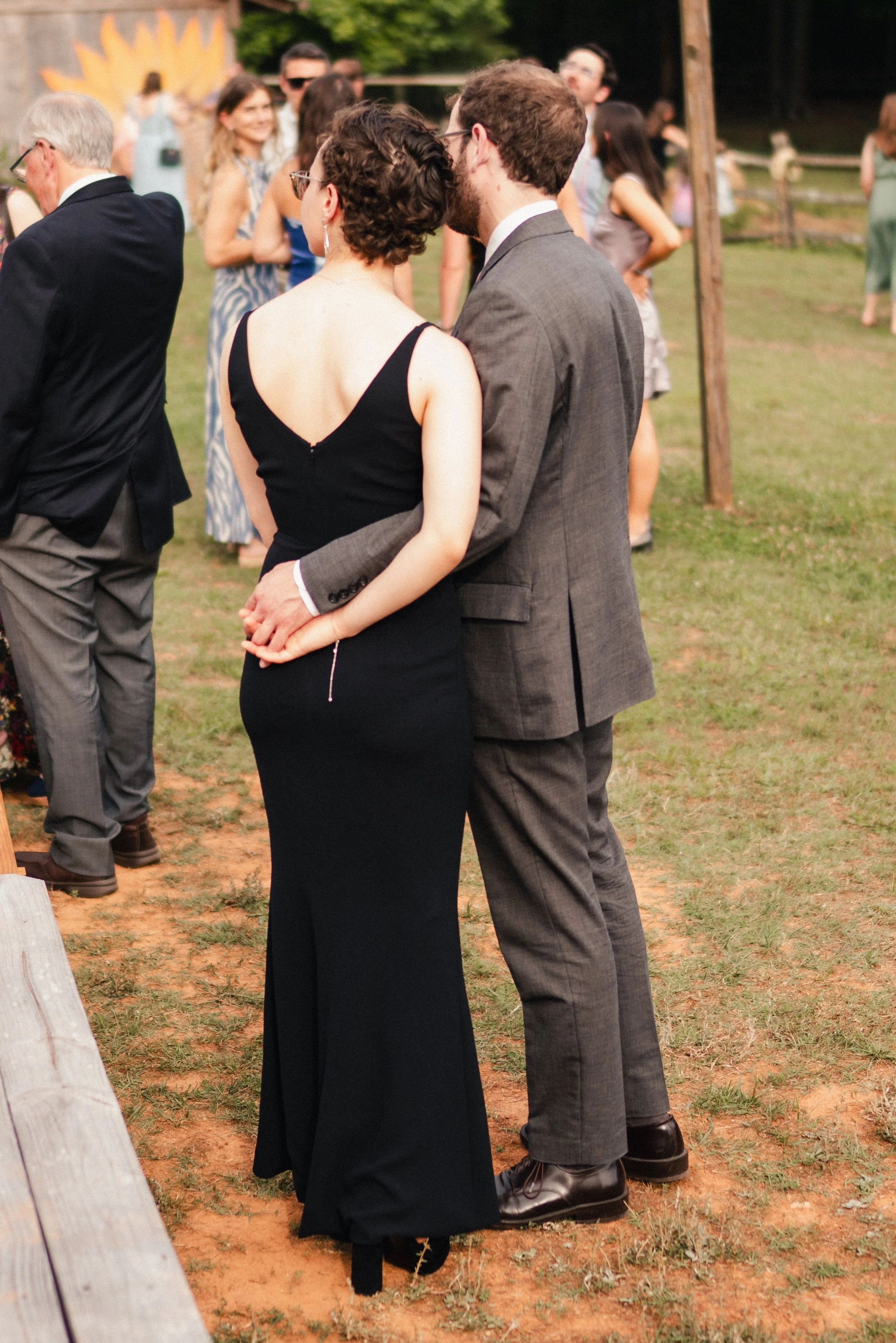 A couple dressed in formal attire, holding hands, standing close together at an outdoor event, with other people socializing in the background.