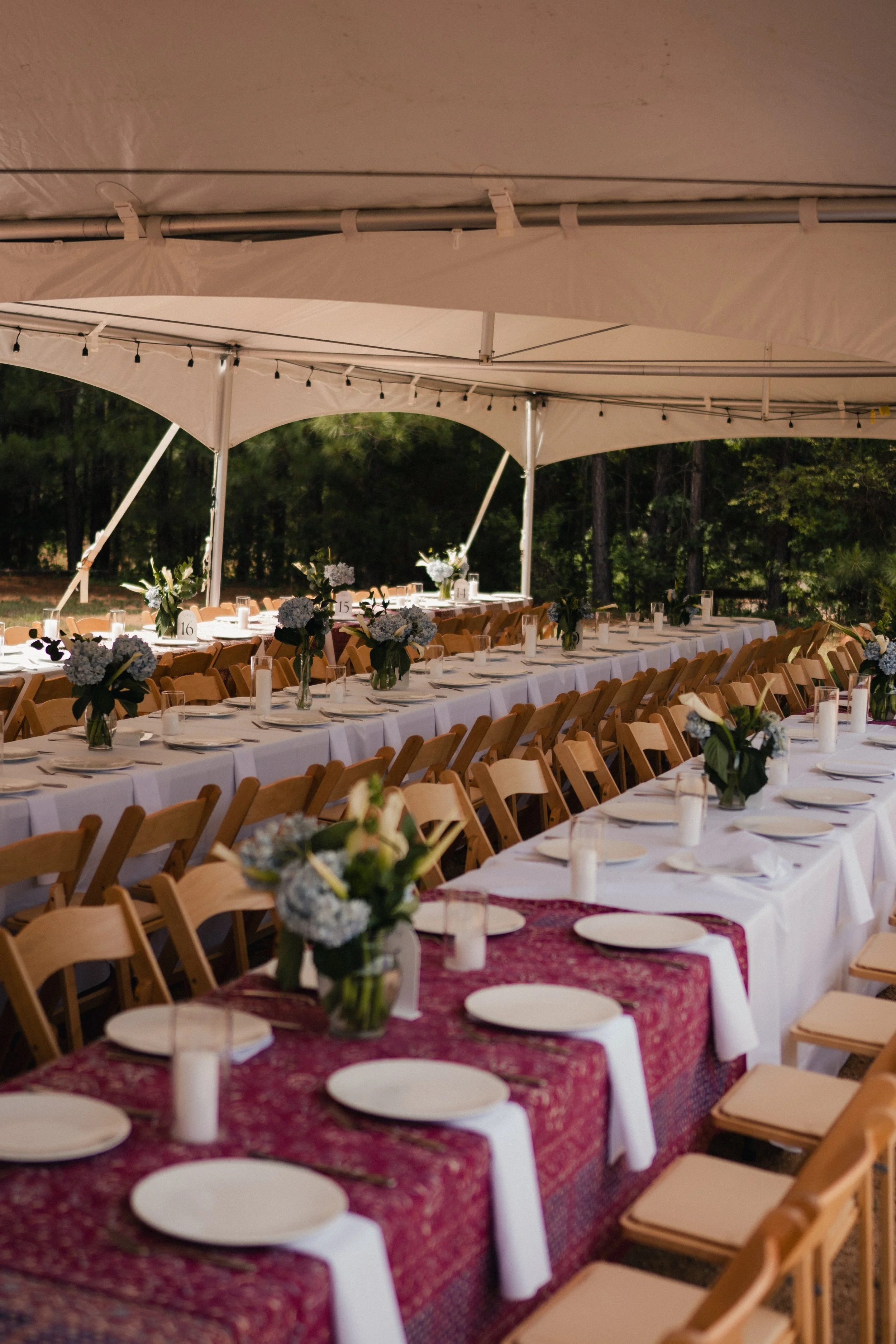 Long banquet table decorated with floral centerpieces under a large white canopy tent, set with white plates, utensils, glasses, and white candles, surrounded by wooden chairs, outdoors in a wooded area at dusk.
