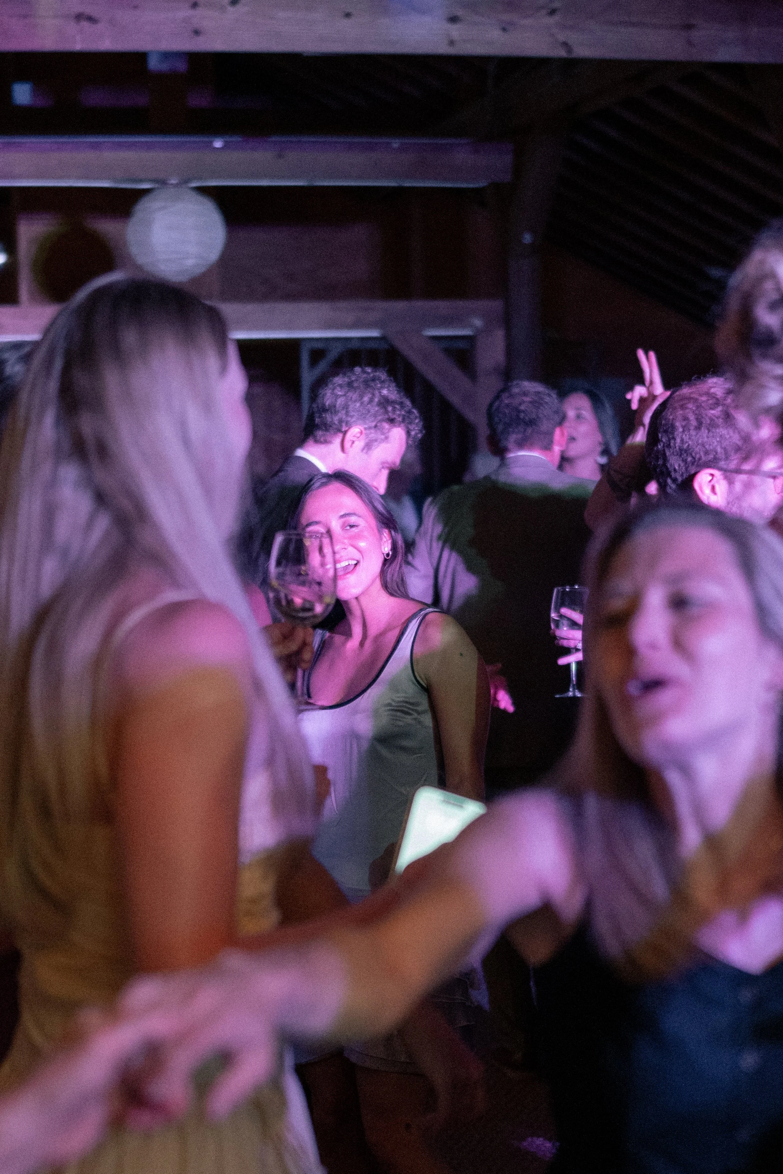 People socializing at a party or event in a dimly lit space with purple lighting, with some holding wine glasses.