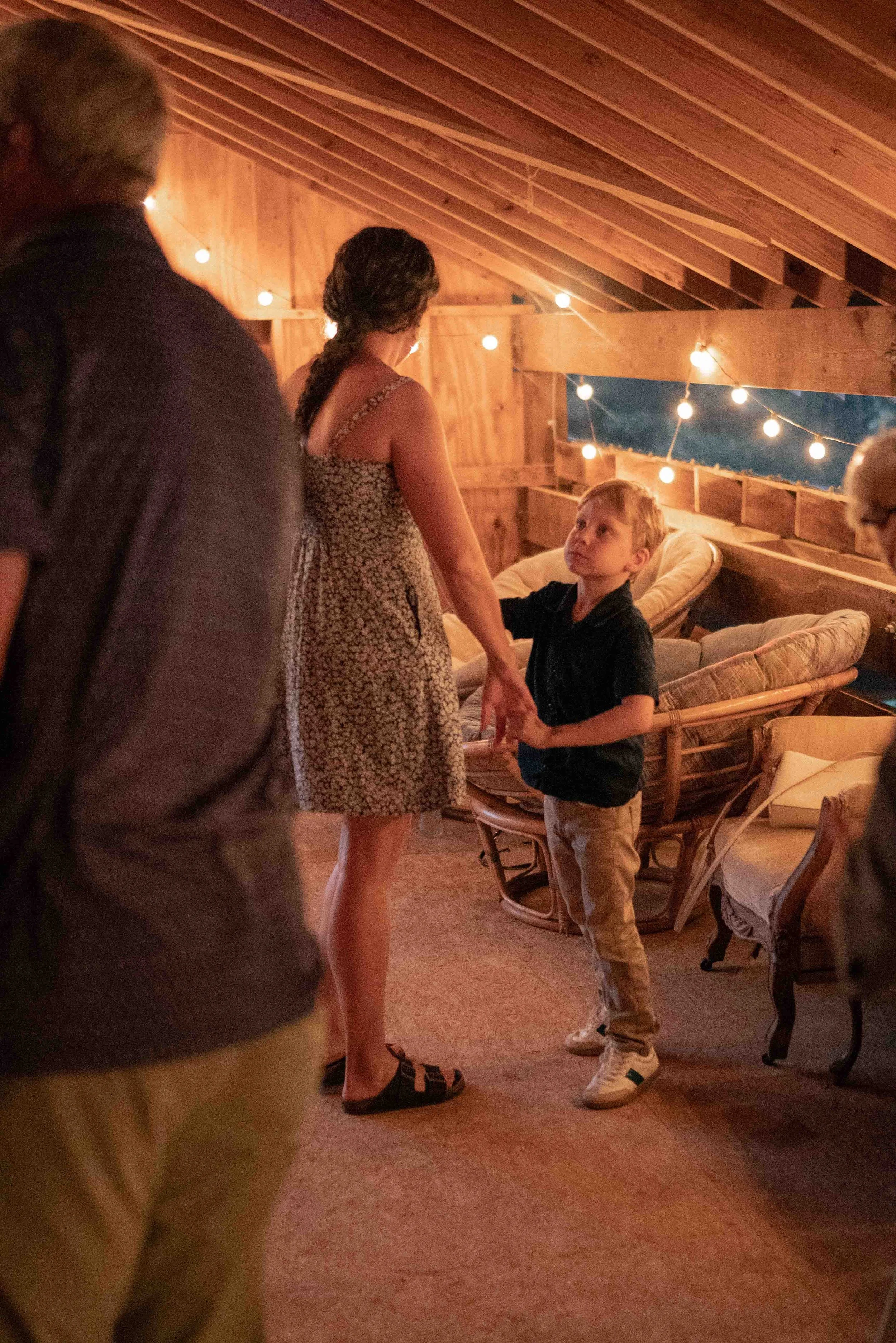 People dancing at a gathering in a wooden room with string lights, including a woman with braided hair and a black dress, and a boy with blond hair and dark shirt, holding hands and looking at each other.
