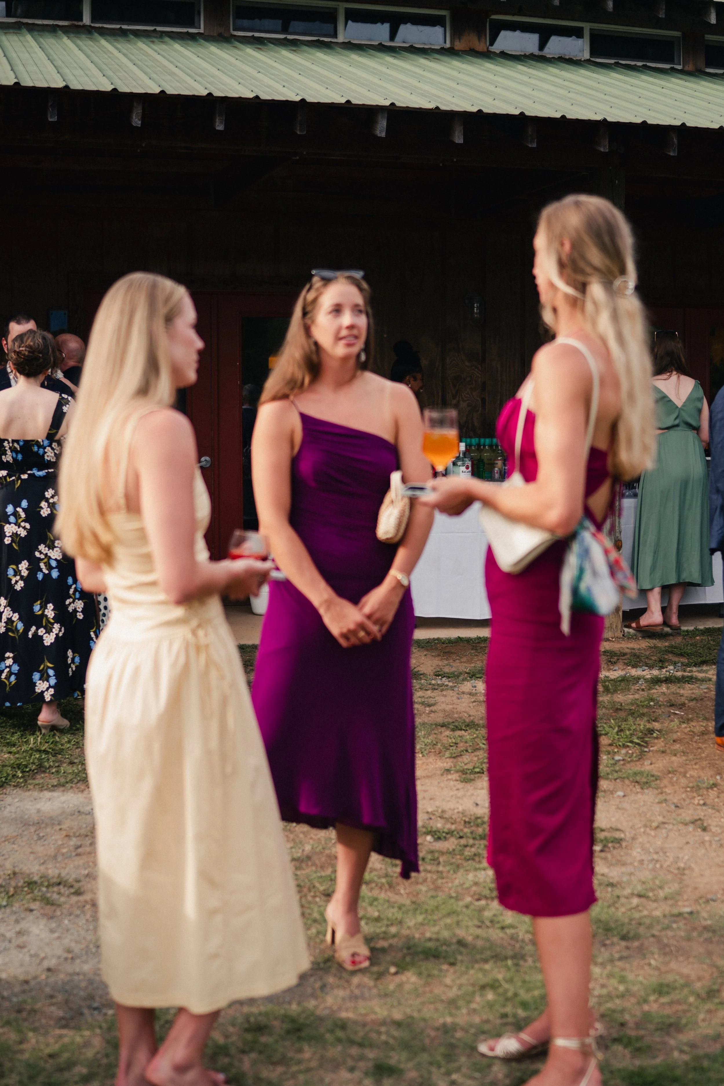 Three women standing outside, engaged in conversation, with drinks in hand, during a social event.
