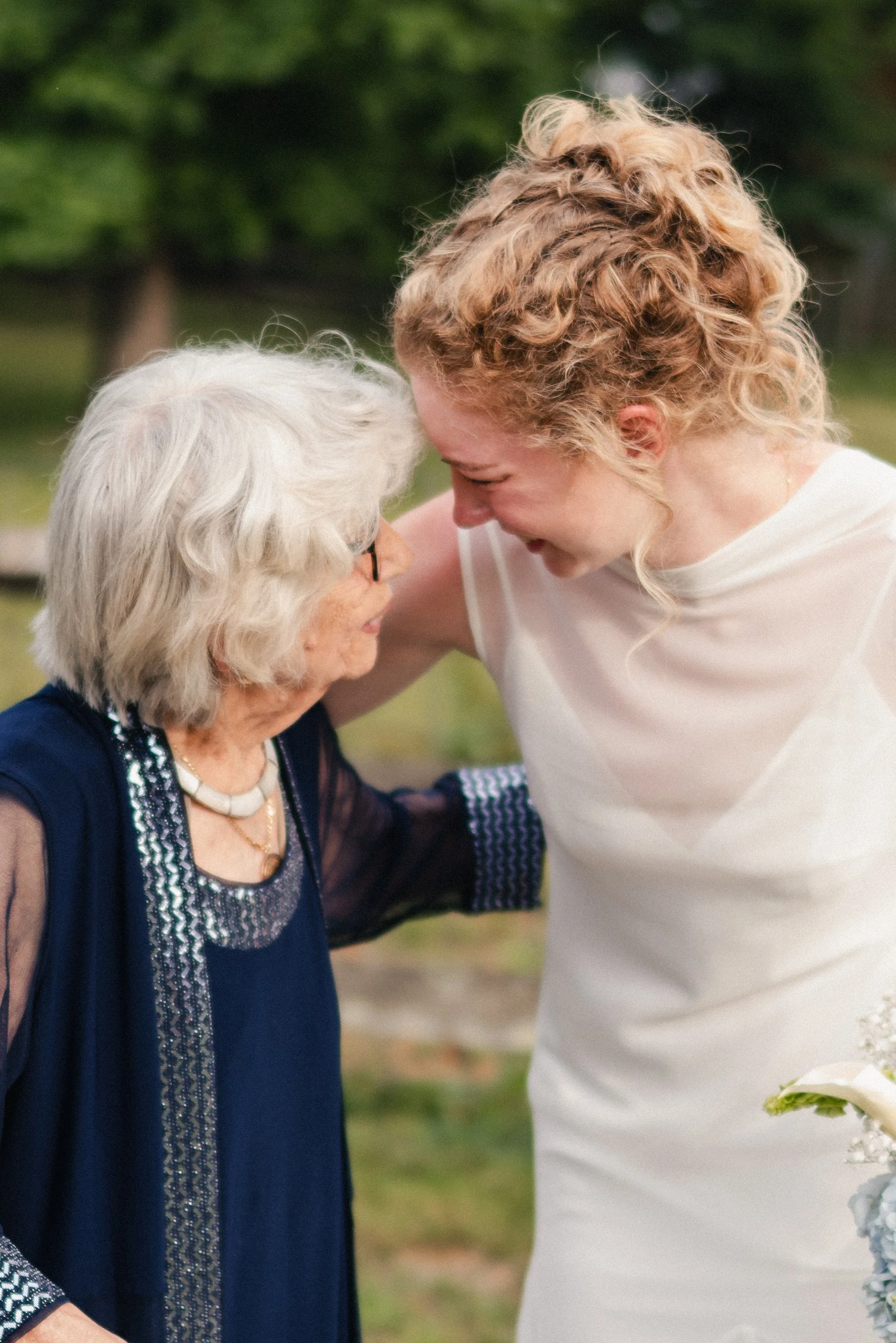 A young woman with curly blonde hair leaning her forehead against an elderly woman with white hair, both smiling and appearing happy outdoors.