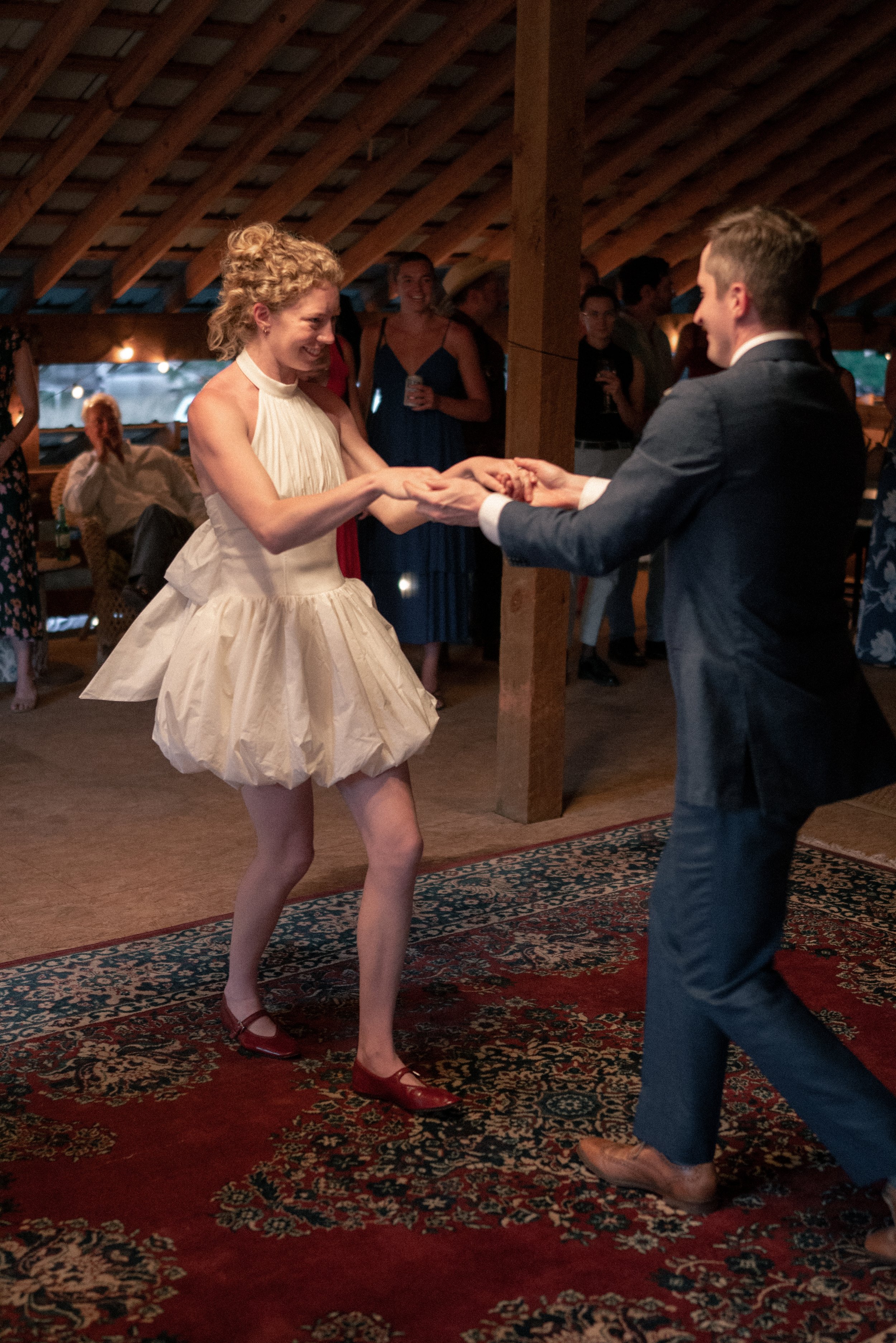 A woman in a cream-colored dress and red shoes dancing with a man in a suit during a wedding reception under a wooden ceiling with a patterned rug on the floor.