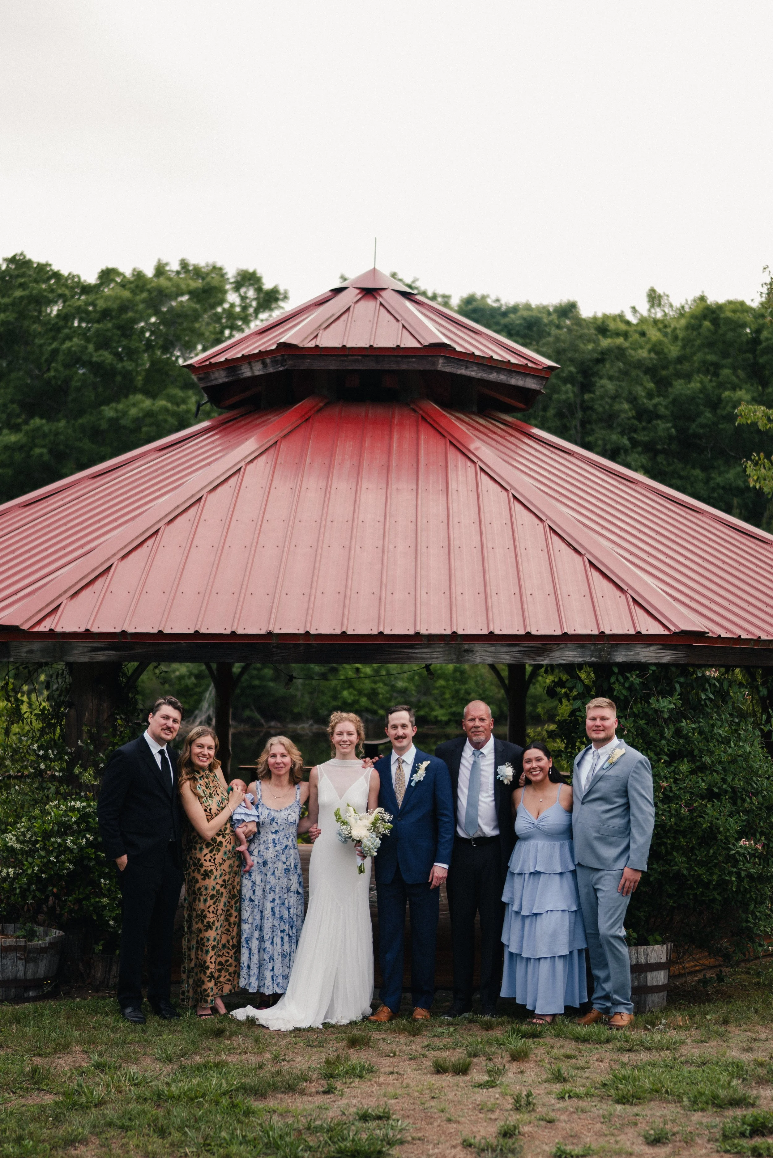 A group of ten people gathered together outdoors under a red metal gazebo roof in a garden setting, with trees in the background, celebrating a wedding. The bride is in a white dress holding a bouquet, and the groom is next to her in a blue suit. Fam