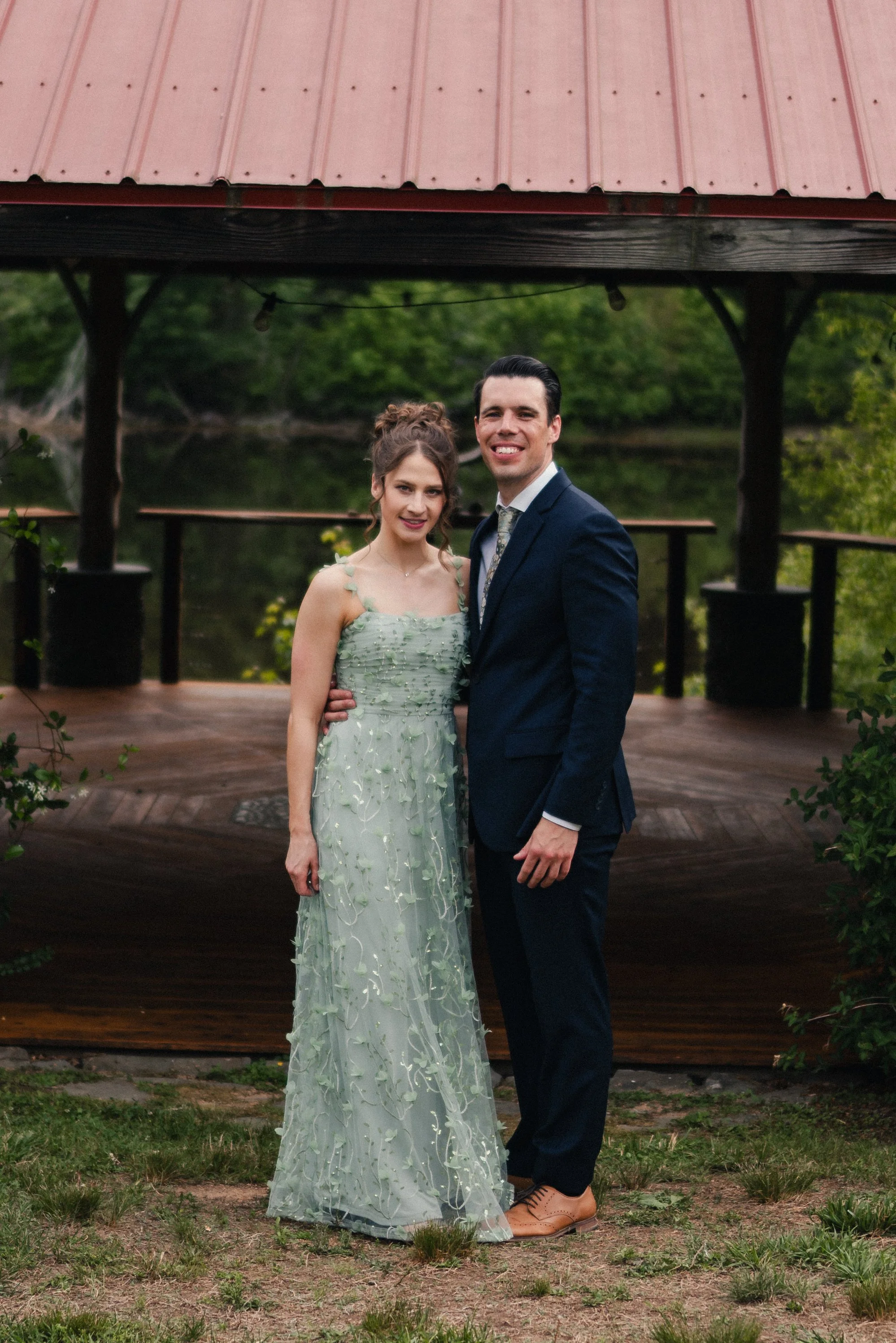 A man and woman dressed in formal attire, standing outdoors in front of a small wooden structure with a red metal roof, surrounded by greenery.
