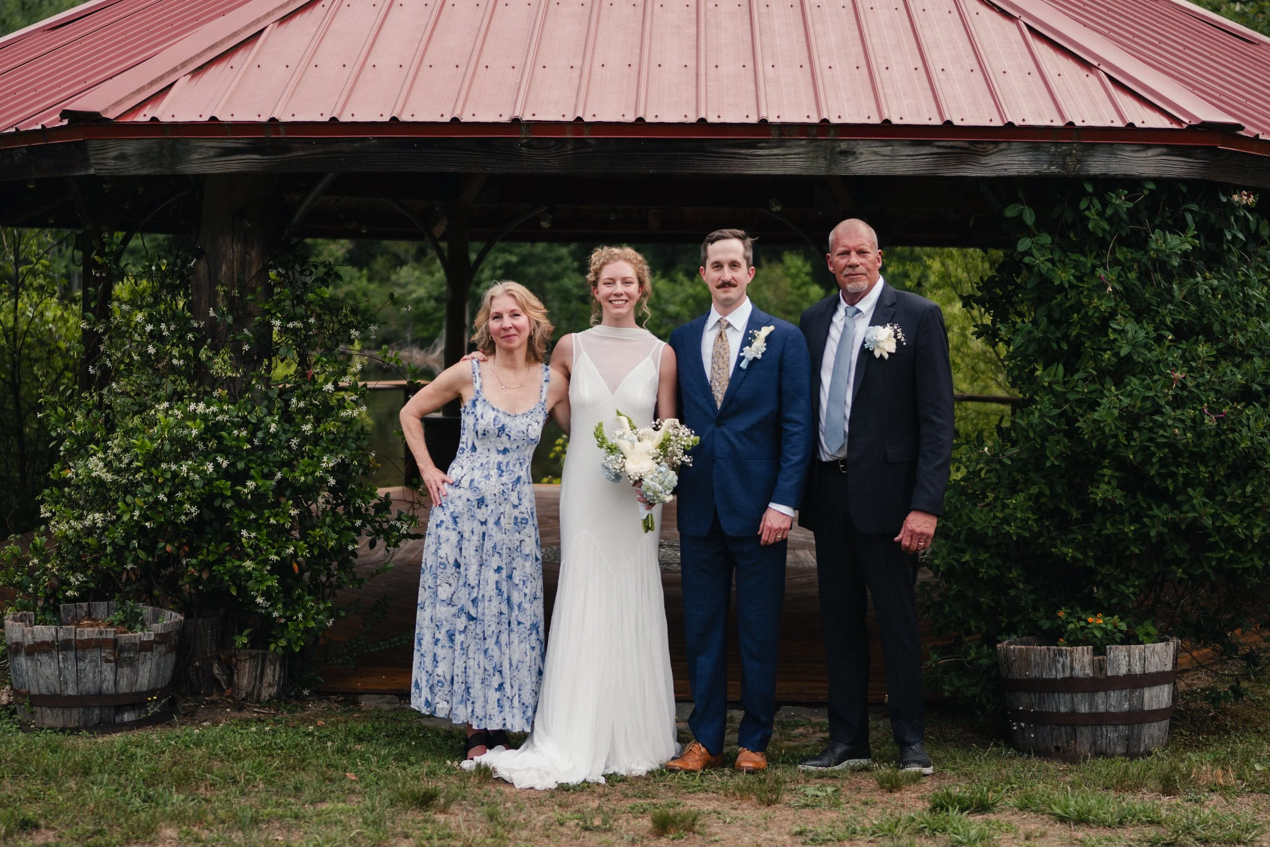 Four people standing together outdoors, with a red metal roofed structure and greenery in the background. The woman in the center is wearing a white wedding dress, holding a bouquet, indicating a wedding celebration.