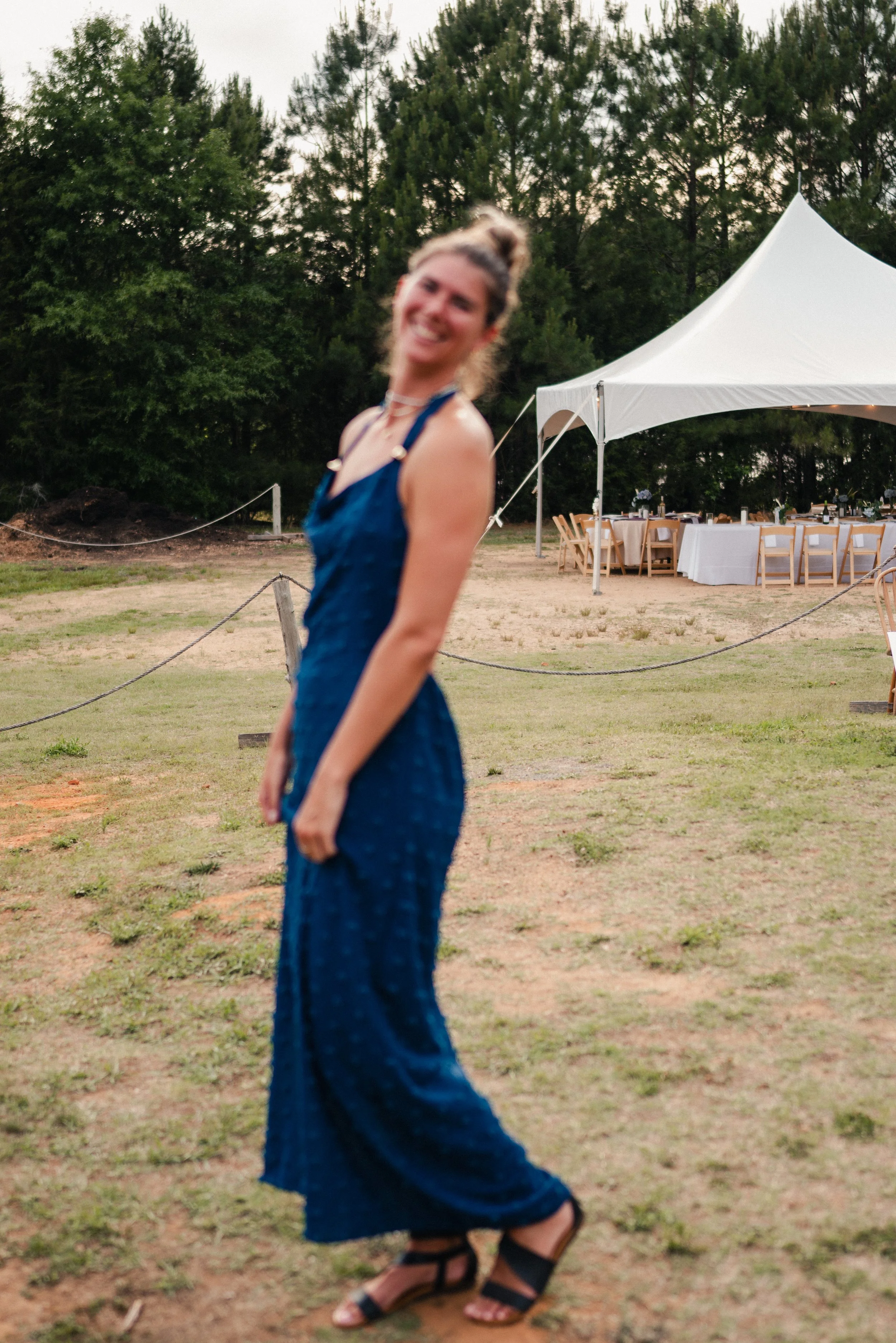 A woman in a blue dress standing outdoors near a white event tent with tables set for an event, surrounded by green trees.